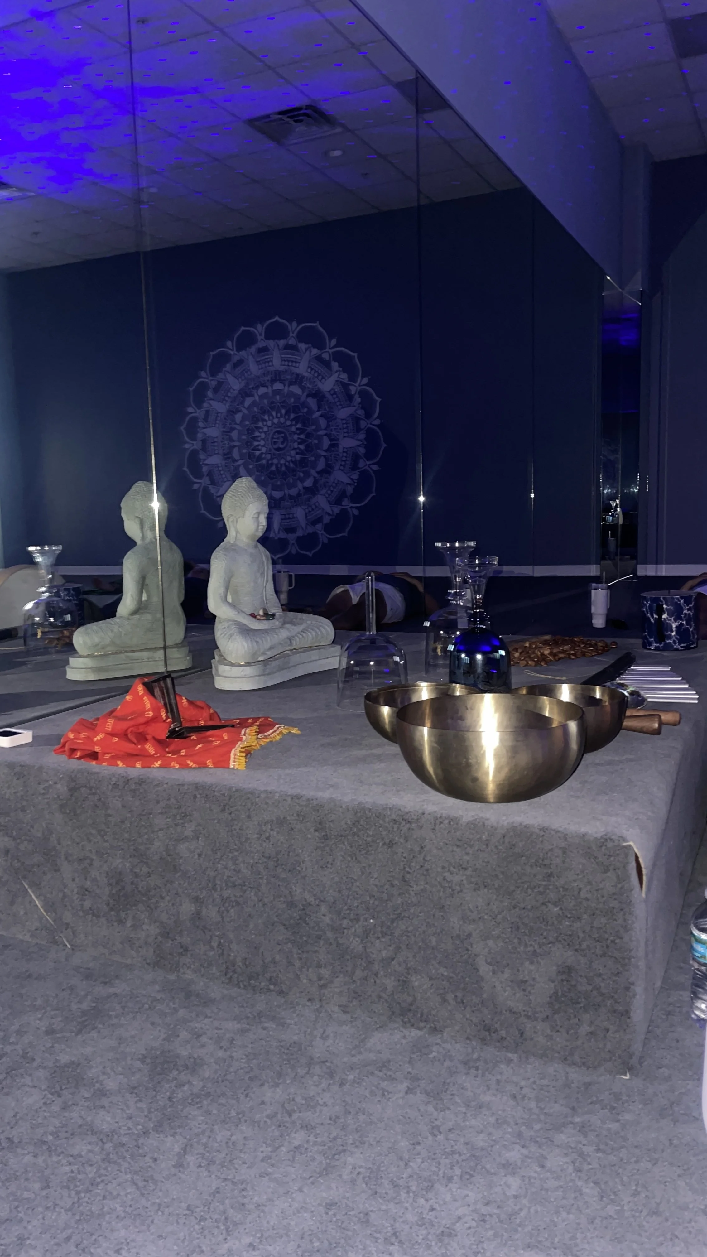 A table decorated with two small statues of meditating children, several wine glasses, metallic bowls, candles, and various utensils, set against a mirrored wall reflecting a blue and purple mandala design.