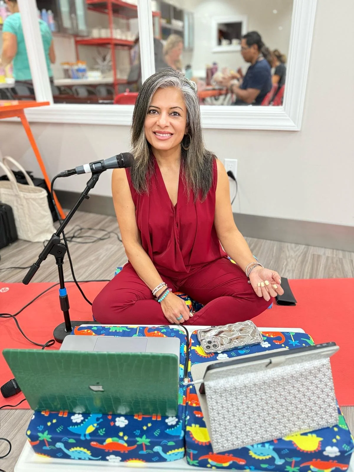 Woman sitting cross-legged at a table with a microphone, laptop, tablet, phone, and open cloth-covered storage box in front of her, smiling at the camera in a room with other people in the background.