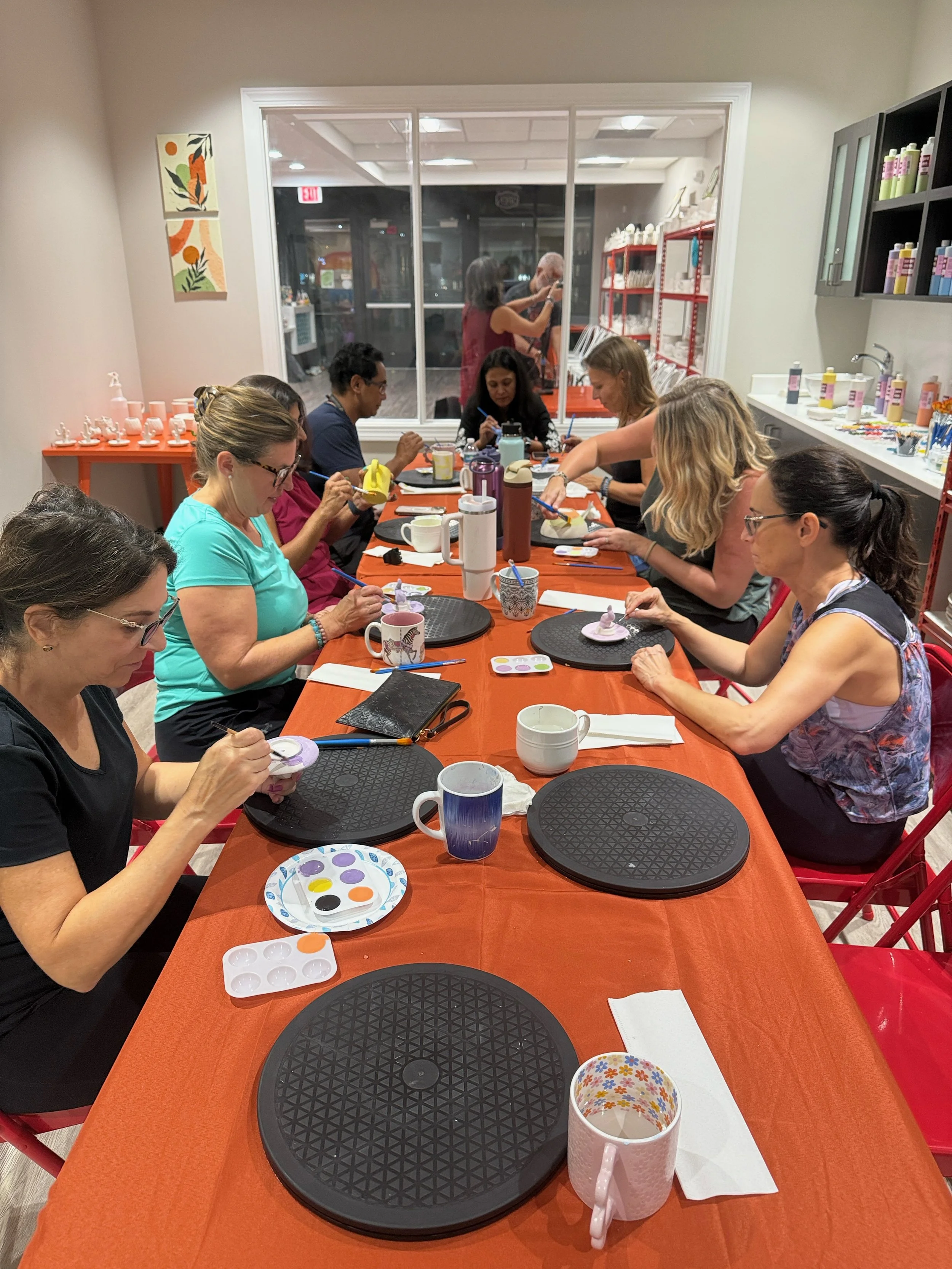 A group of people sitting around a long orange table, painting small ceramic objects in a craft workshop. There are art supplies like paints and brushes on the table, and shelves with bottles on the wall.
