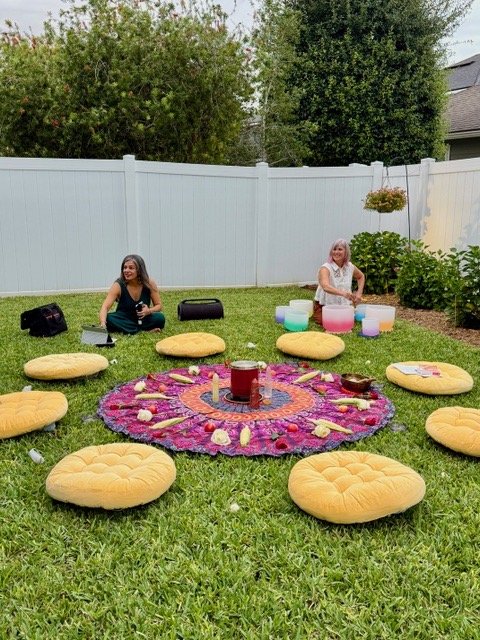 Two young girls in a backyard, sitting on the grass near a white fence. One girl is seated with a black bag next to her, and the other girl is smiling and near colorful singing bowls. Cushions are arranged in a circle around a vibrant, decorated rug with candles and small objects on it.