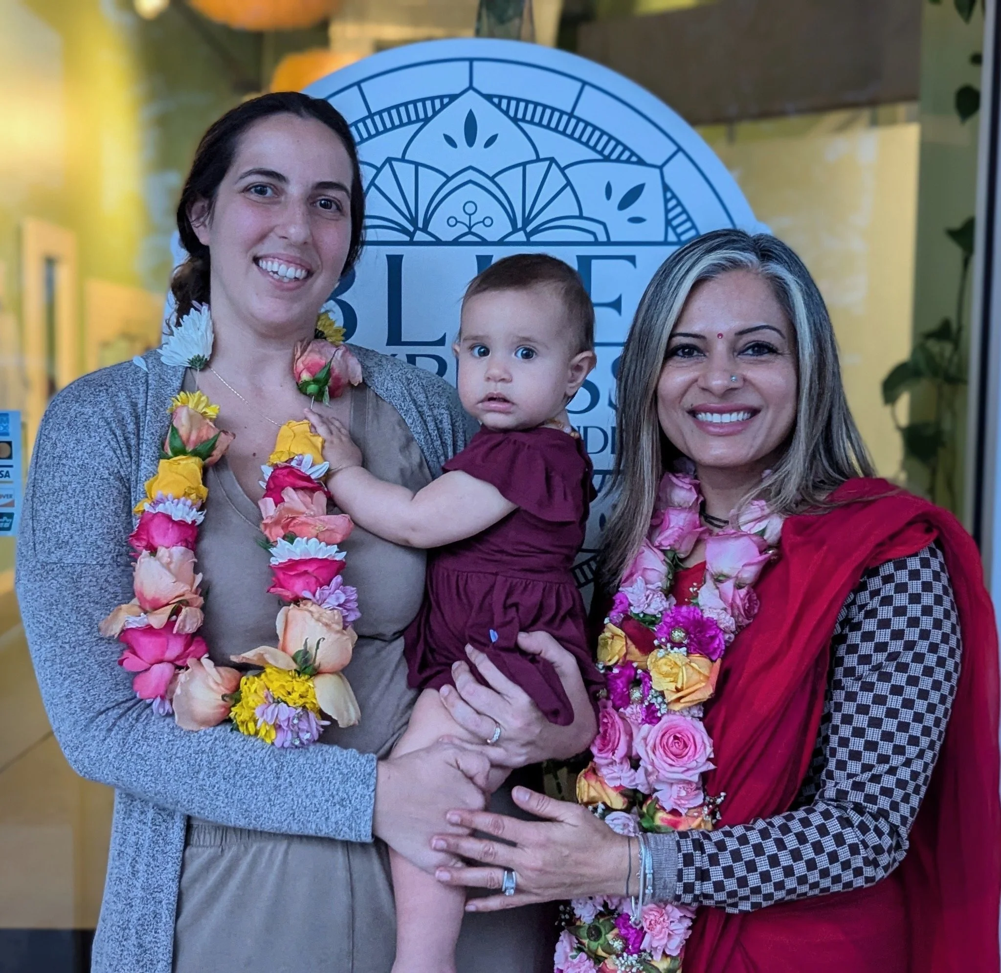 Two women holding a young girl, all wearing flower garlands, in front of a blue and white patterned backdrop with a sign.