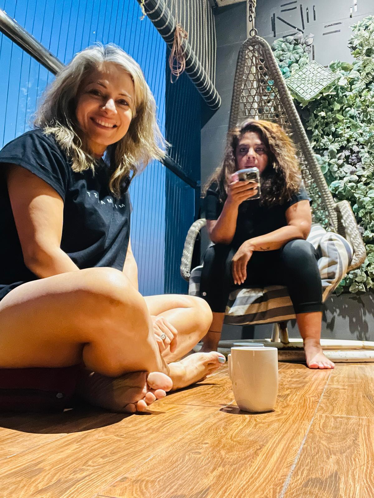 Two women sitting on a wooden floor, one smiling at the camera and the other drinking from a glass, with a coffee mug on the floor in front of them, in a cozy indoor setting with plants and hanging chairs.