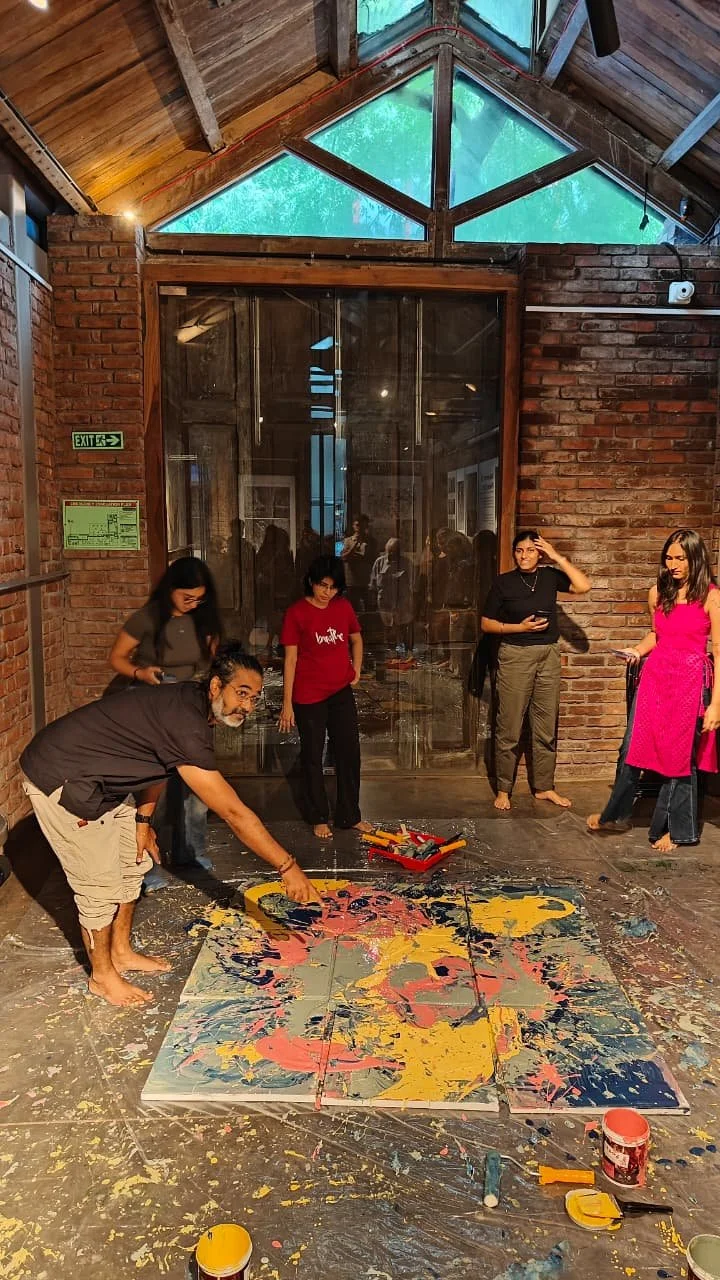 Group of people creating an abstract painting on four canvases using bright colors in an art studio with a brick wall and a glass ceiling.