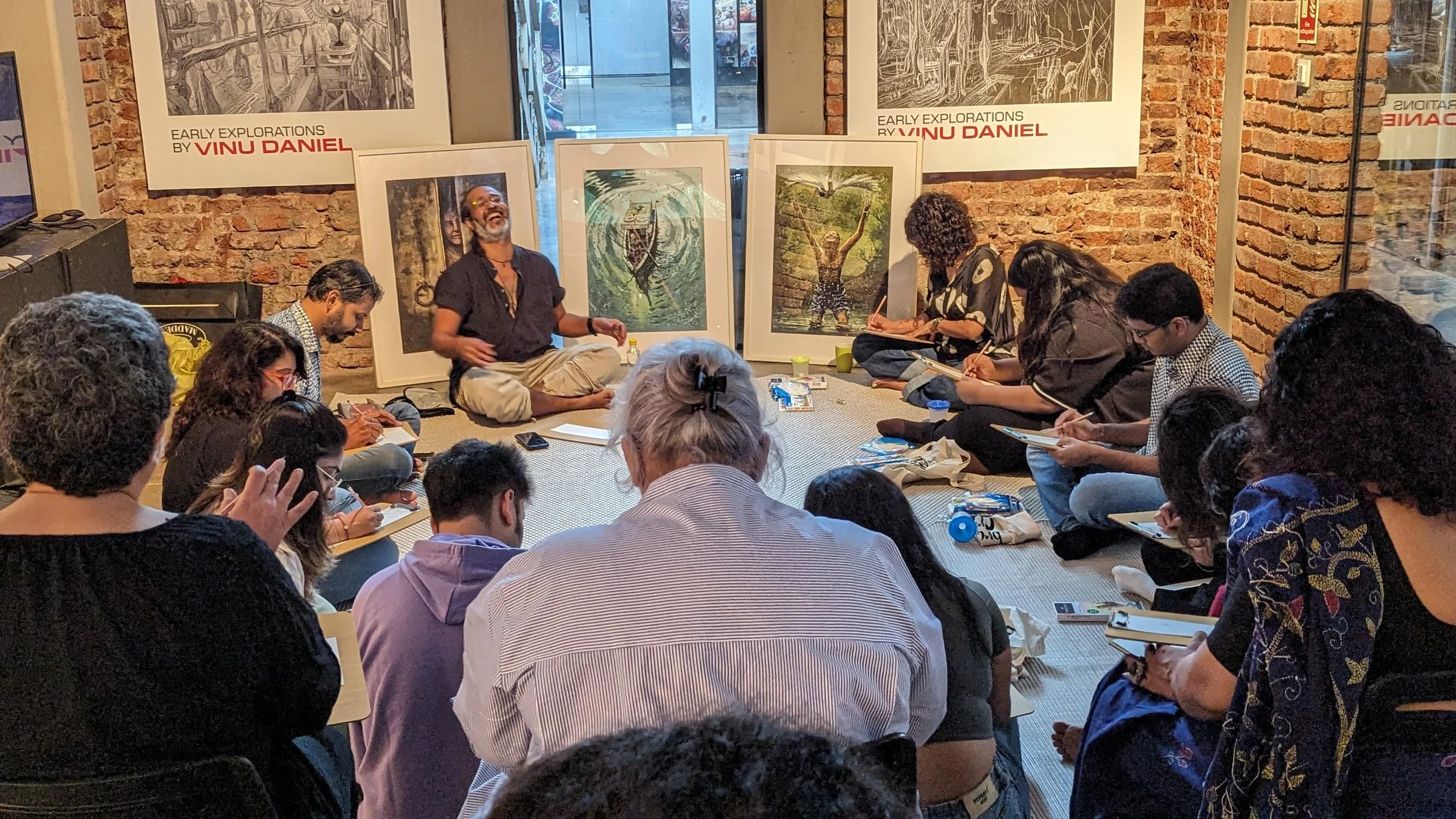 An art talk or workshop with Vinu Daniel seated cross-legged in front of artwork, surrounded by seated audience taking notes, in a gallery with brick walls and window.