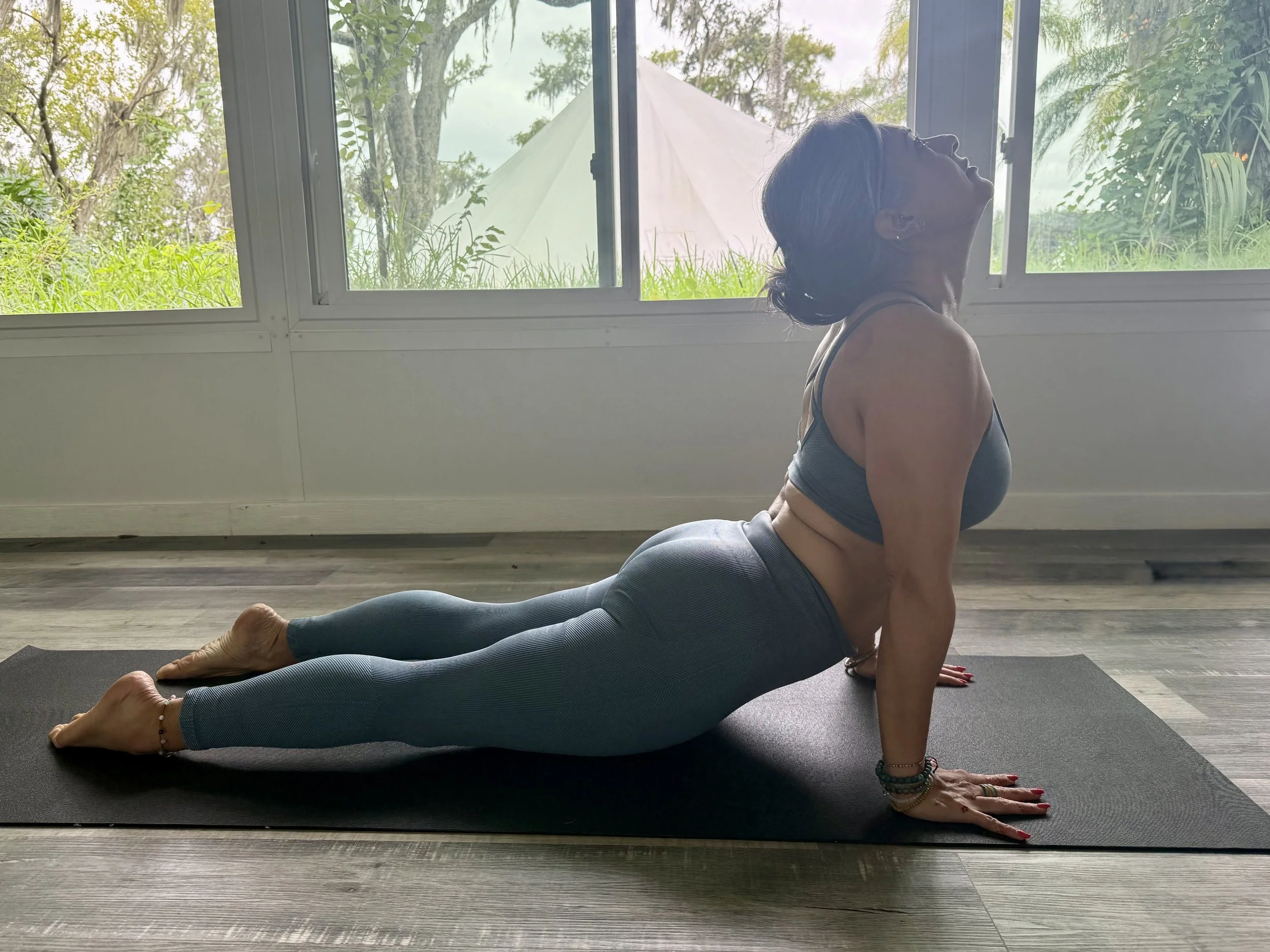 A woman practicing yoga in a yoga pose on a black mat in a room with large windows showing greenery outside.