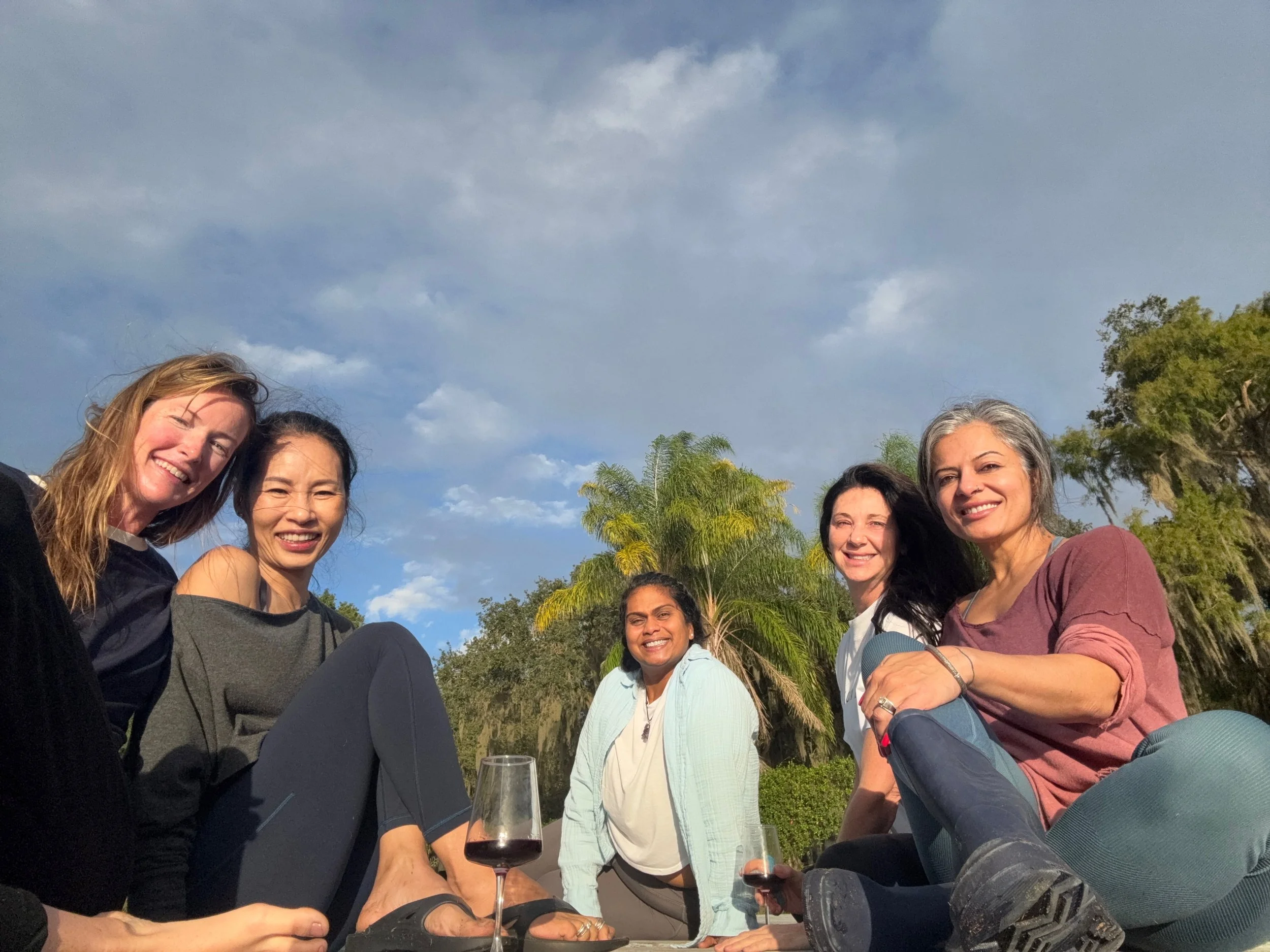 Group of five women sitting outdoors with a blue sky and trees in the background, smiling and enjoying wine.