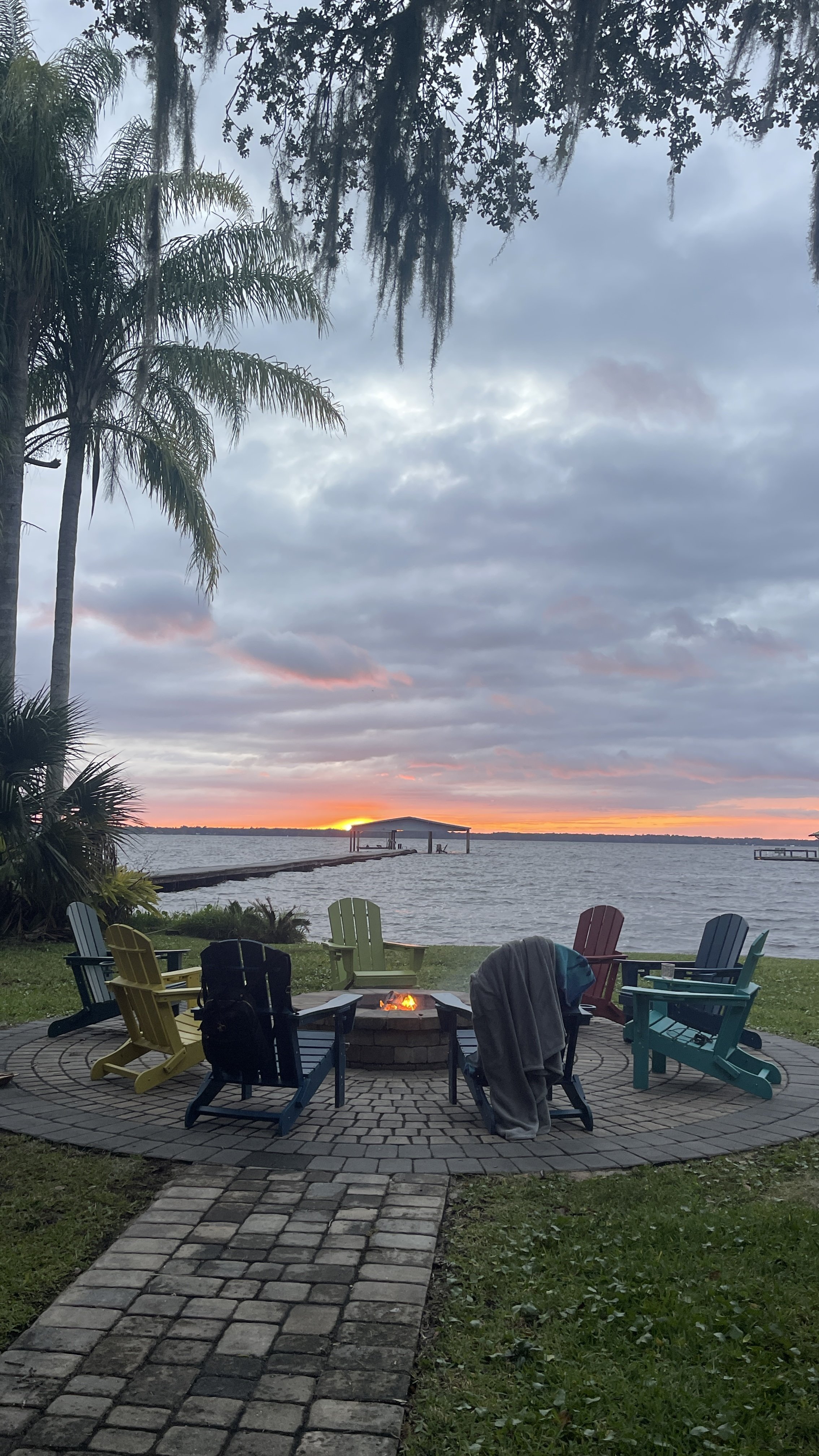 A lakeside scene at sunset with a circular brick fire pit surrounded by colorful Adirondack chairs, some with blankets, on a paved patio with grass, overlooking a lake with a dock and a covered boat slip, under a cloudy sky with hints of sunset colors.