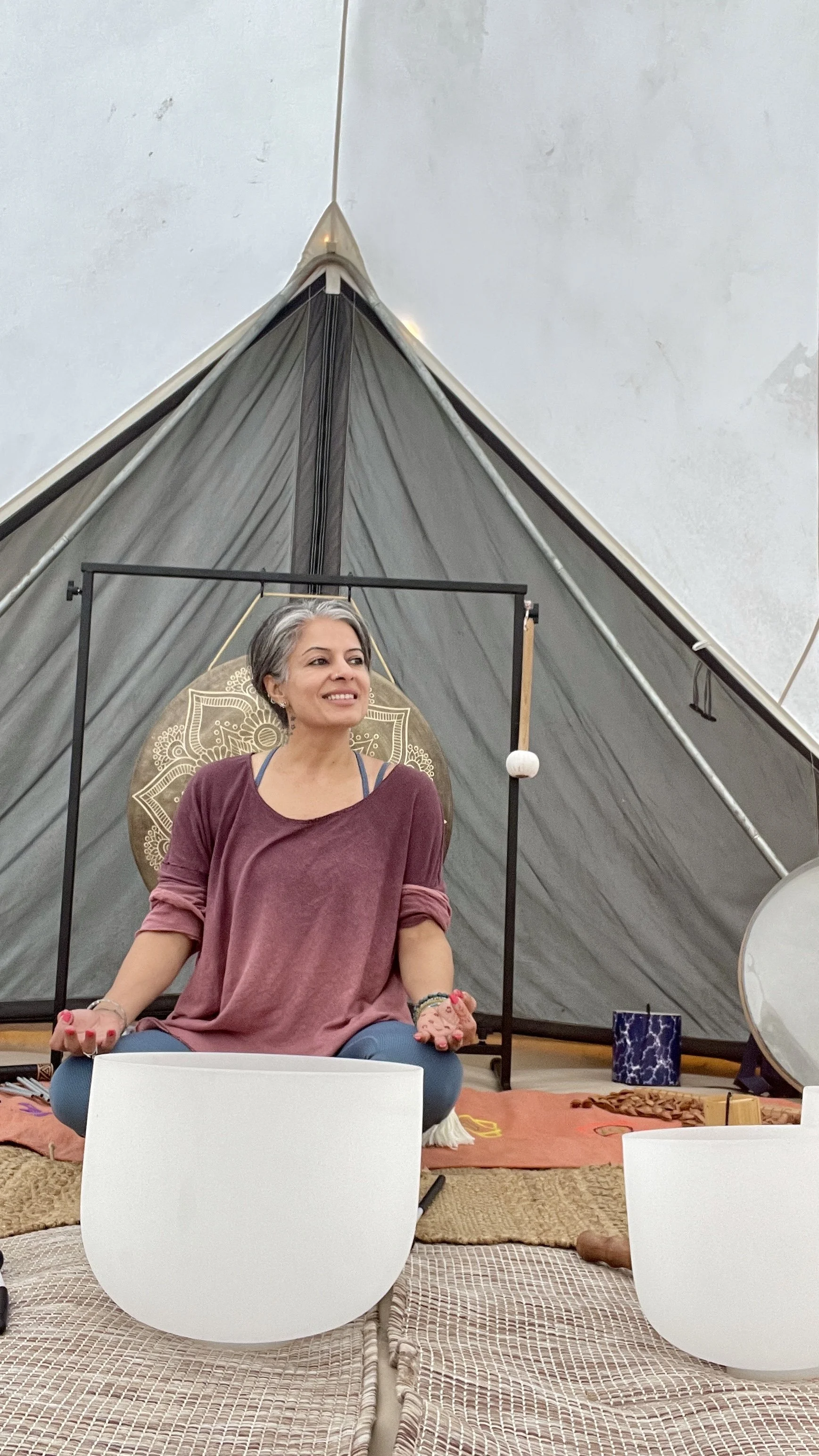 A woman with short gray hair sitting cross-legged inside a tent, surrounded by yoga and meditation props, with a decorative circle behind her, holding her hands in a meditation pose.