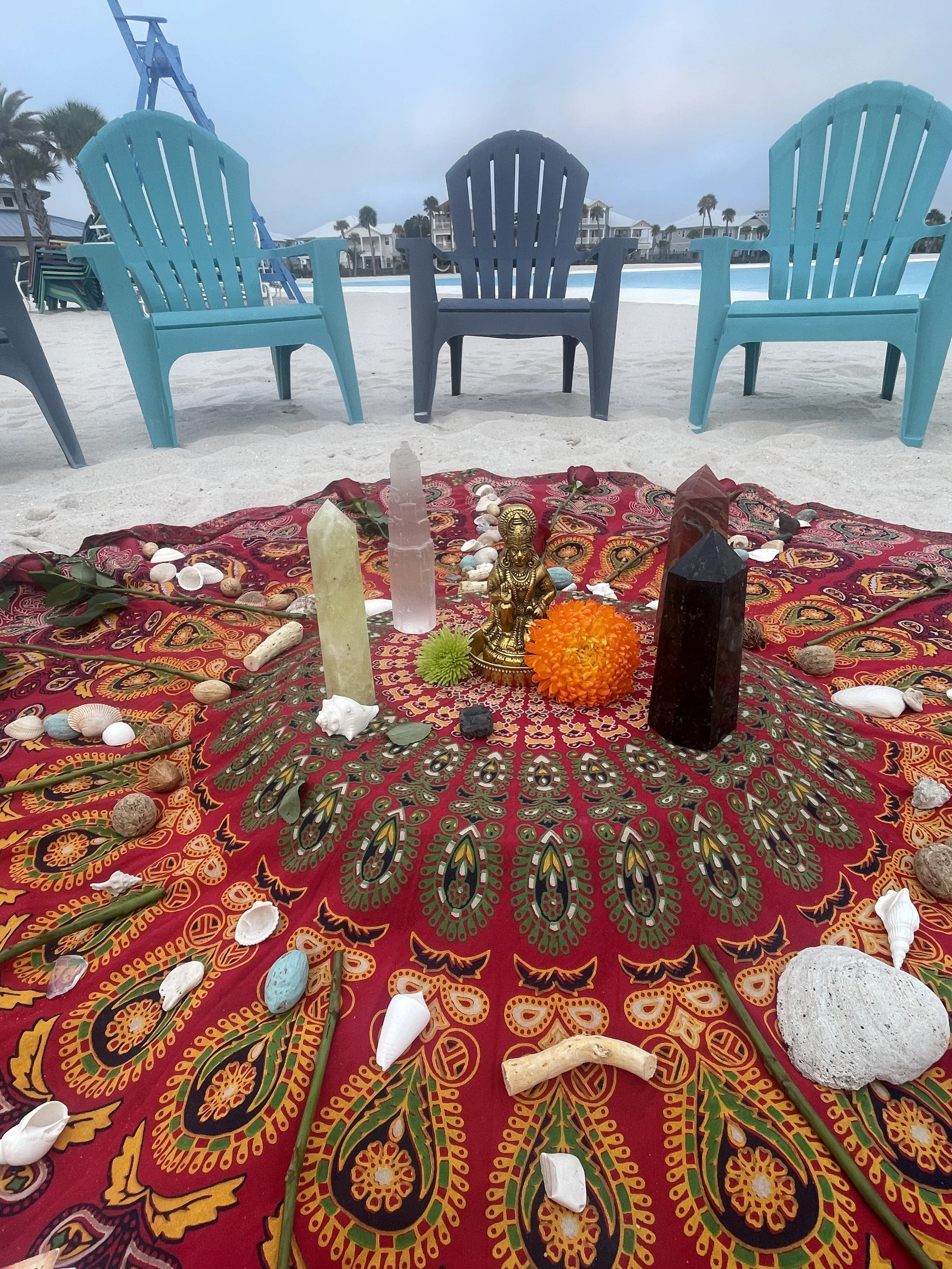 Two young girls in a backyard, sitting on the grass near a white fence. One girl is seated with a black bag next to her, and the other girl is smiling and near colorful singing bowls. Cushions are arranged in a circle around a vibrant, decorated rug with candles and small objects on it.