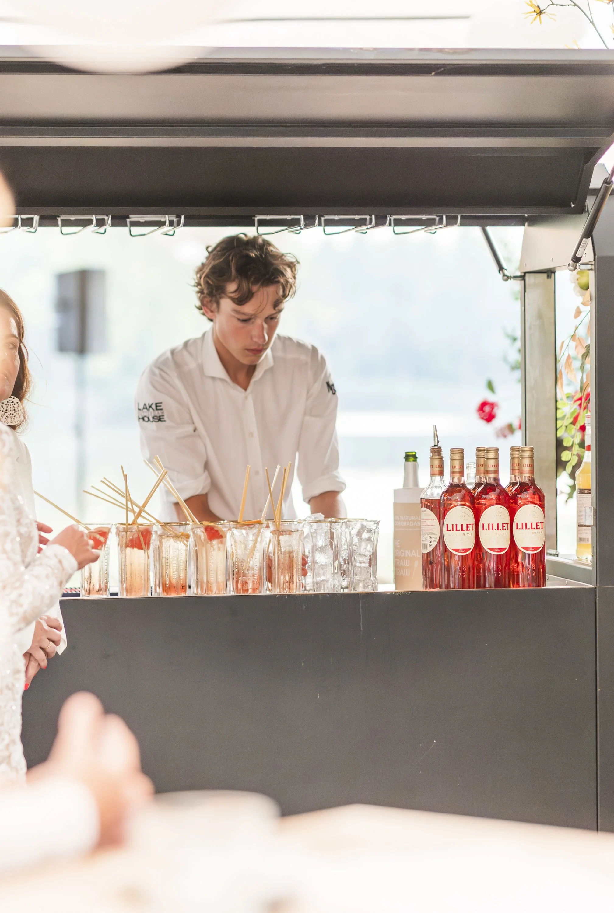een jonge man in een witte blouse die achter een barrette staat met glazen met een roze drankje en stokjes, en flesjes met rosé wijn op de plank ernaast