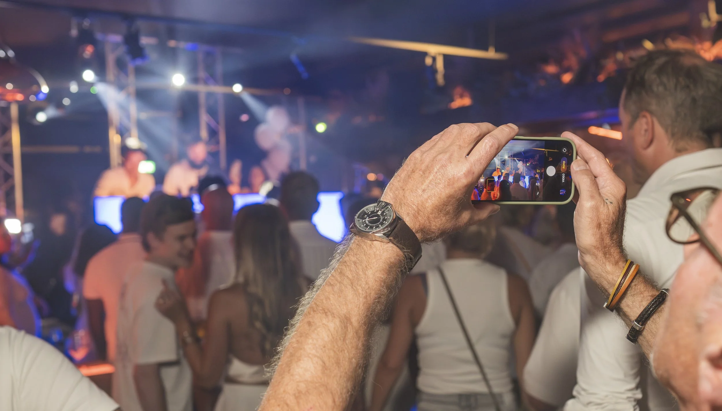 Man houdt een smartphone vast en neemt een foto van een groot publiek en een podium met lichtshows tijdens een concert of evenement in een donkere zaal.