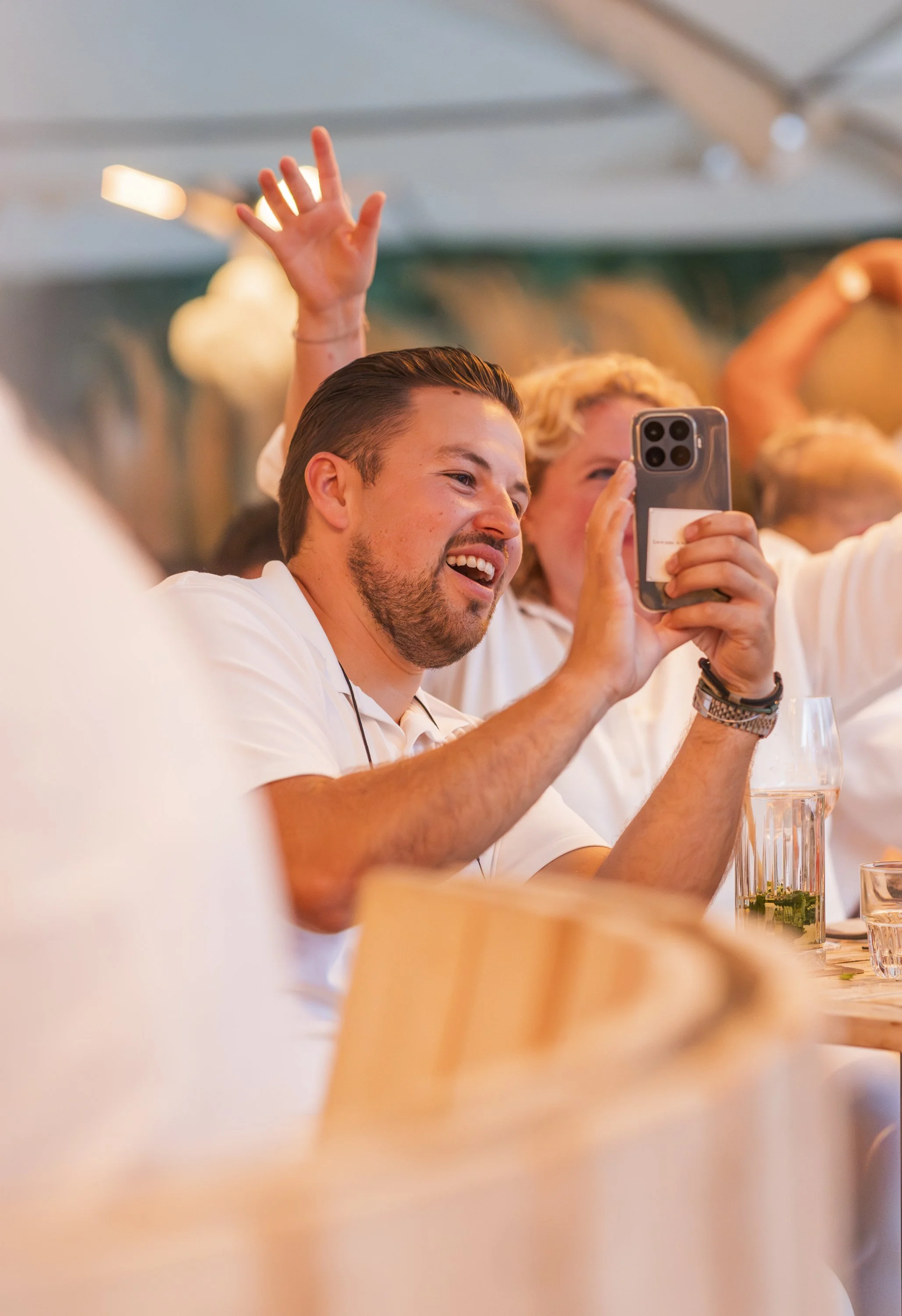 Man die in een restaurant of café met een witte polo aan het eten is. Hij houdt een smartphone vast en is in een groepsactiviteit, anderen heffen handen op en lachen.