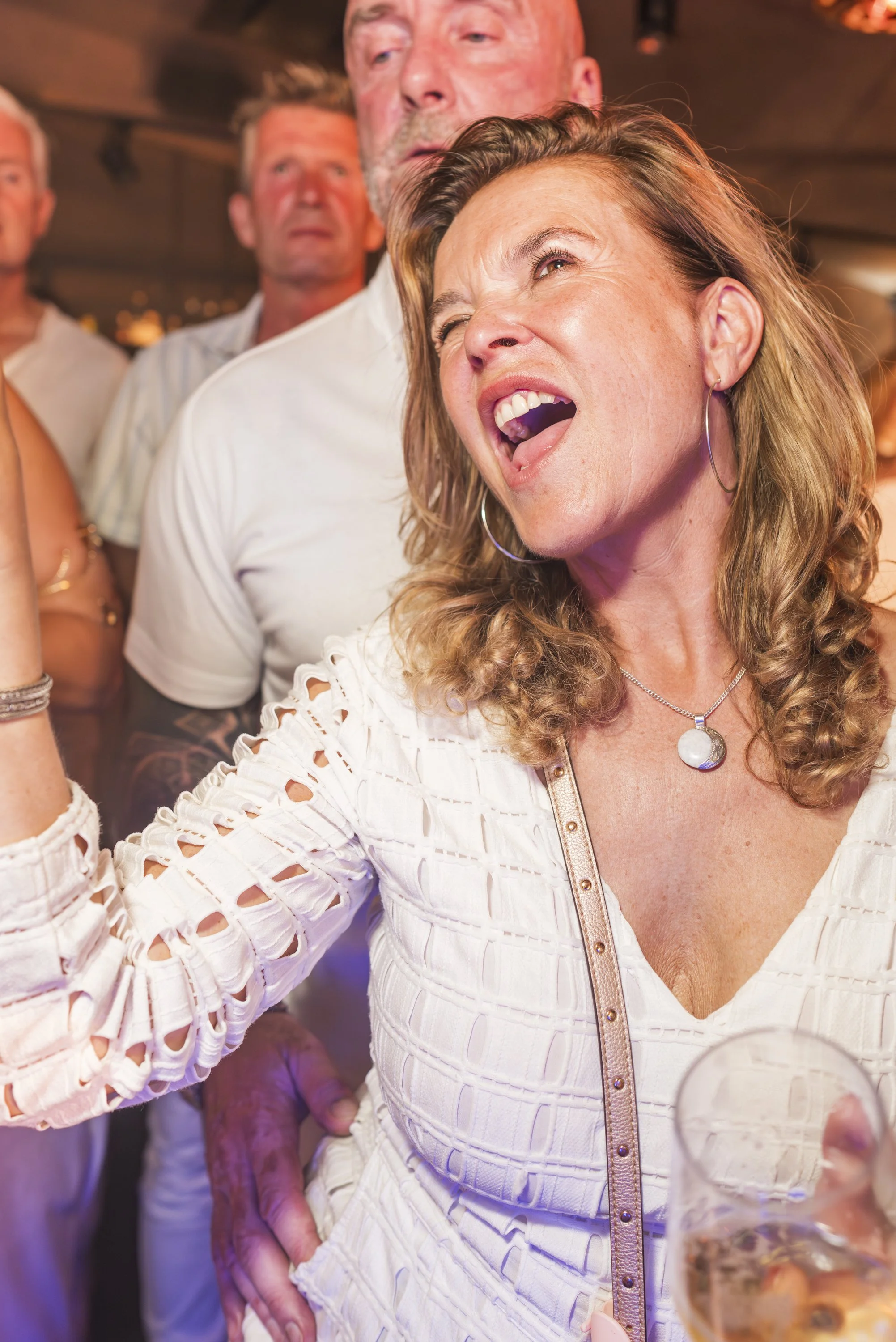 Een vrouw met golvend, lichtbruin haar, grote oorbellen en een witte jurk met wijde mouwen die met openingen zijn versierd, houdt een glas met een drankje in een drukke sociale setting.