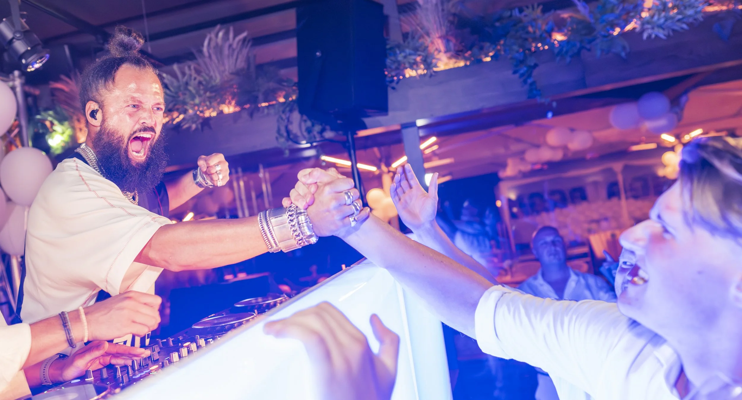 Dansende DJ met lang haar in een hoge paardenstaart en een beard, high-fiving met een vrouwelijke danser in een kleurrijke setting met neonlichten en planten.