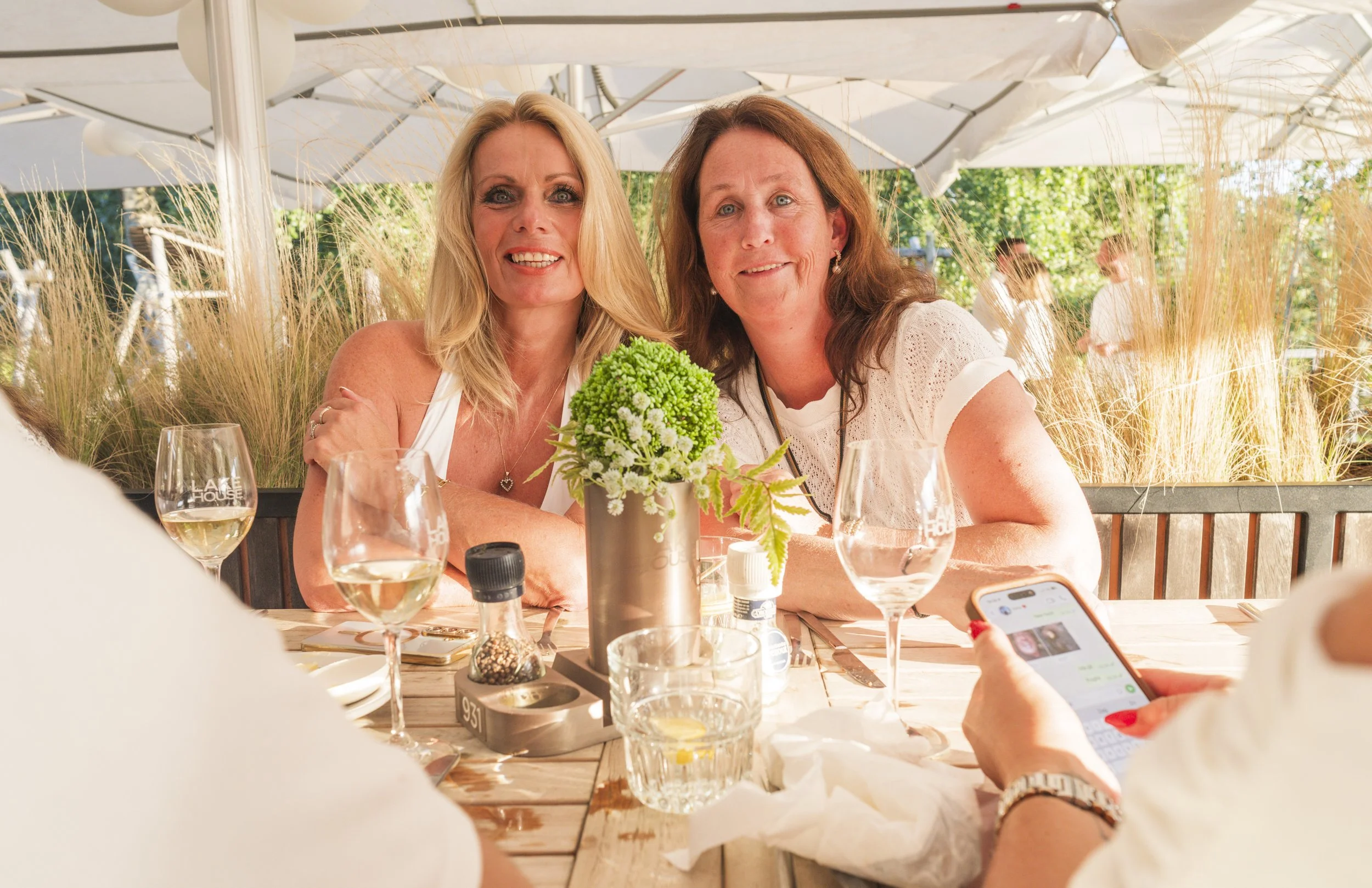 Twee vrouwen zitten samen aan een tafel in een restaurant, omringd door glazen wijn, bloemen en allerlei eten, onder een parasol buiten.