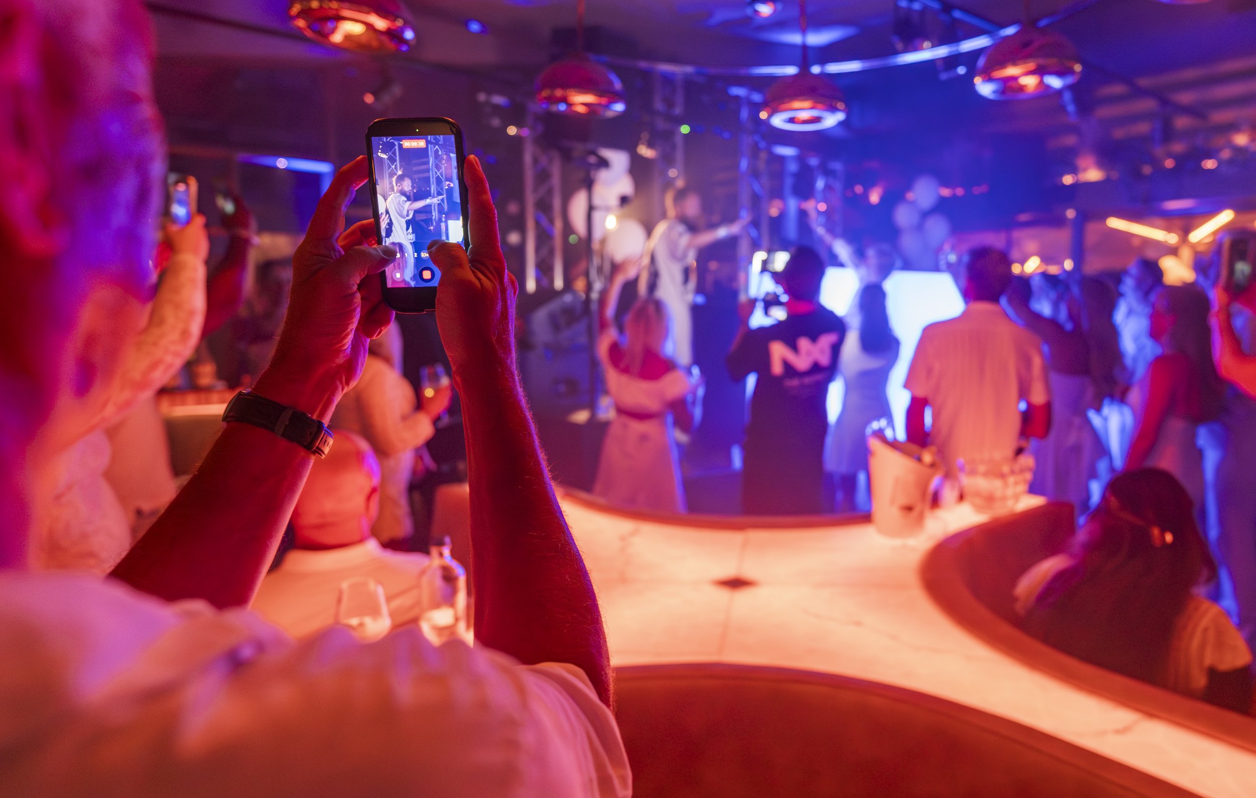 Slecht verlichte dansvloer met mensen die dansen en een live band op het podium, gekleed in feestelijke kleding, in een club of concertzaal met kleurrijk licht.