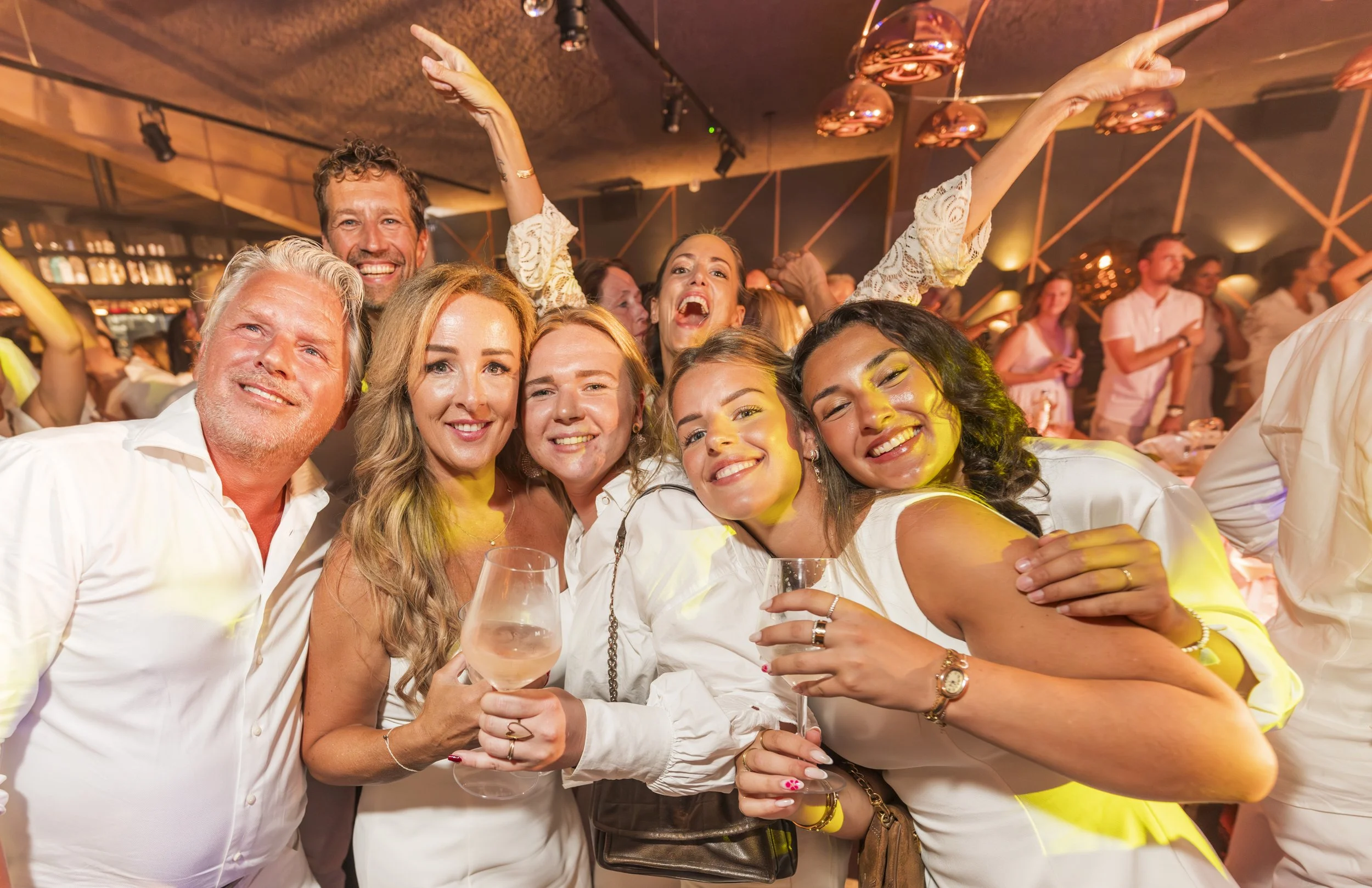 Groepsfoto van feestende mensen, lachend en dansend in een warme sfeer, met een bar op de achtergrond en warme verlichting.