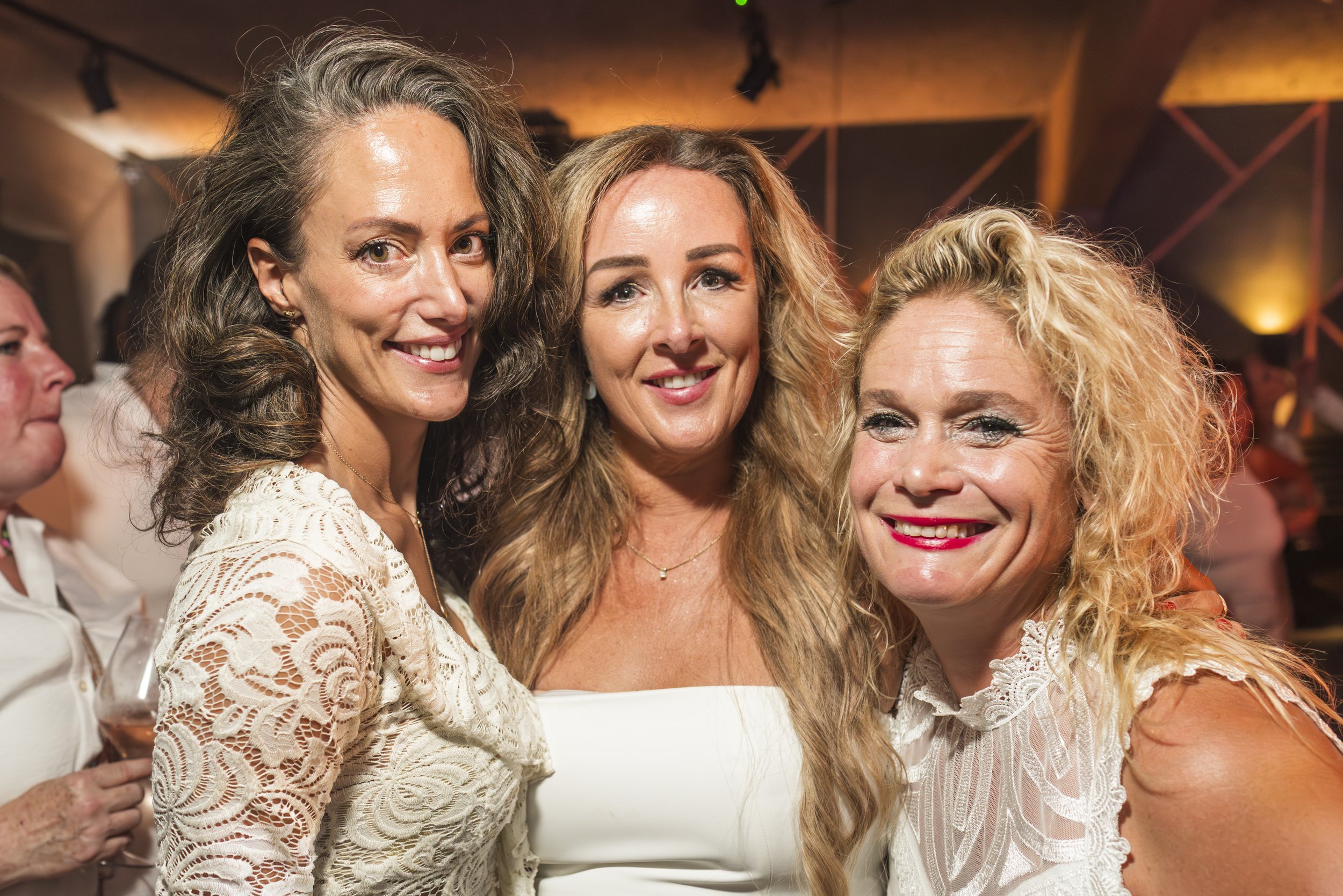 Drie vrouwen die samen poseren en lachen op een feest of evenement, met achtergrondverlichting en andere gasten.