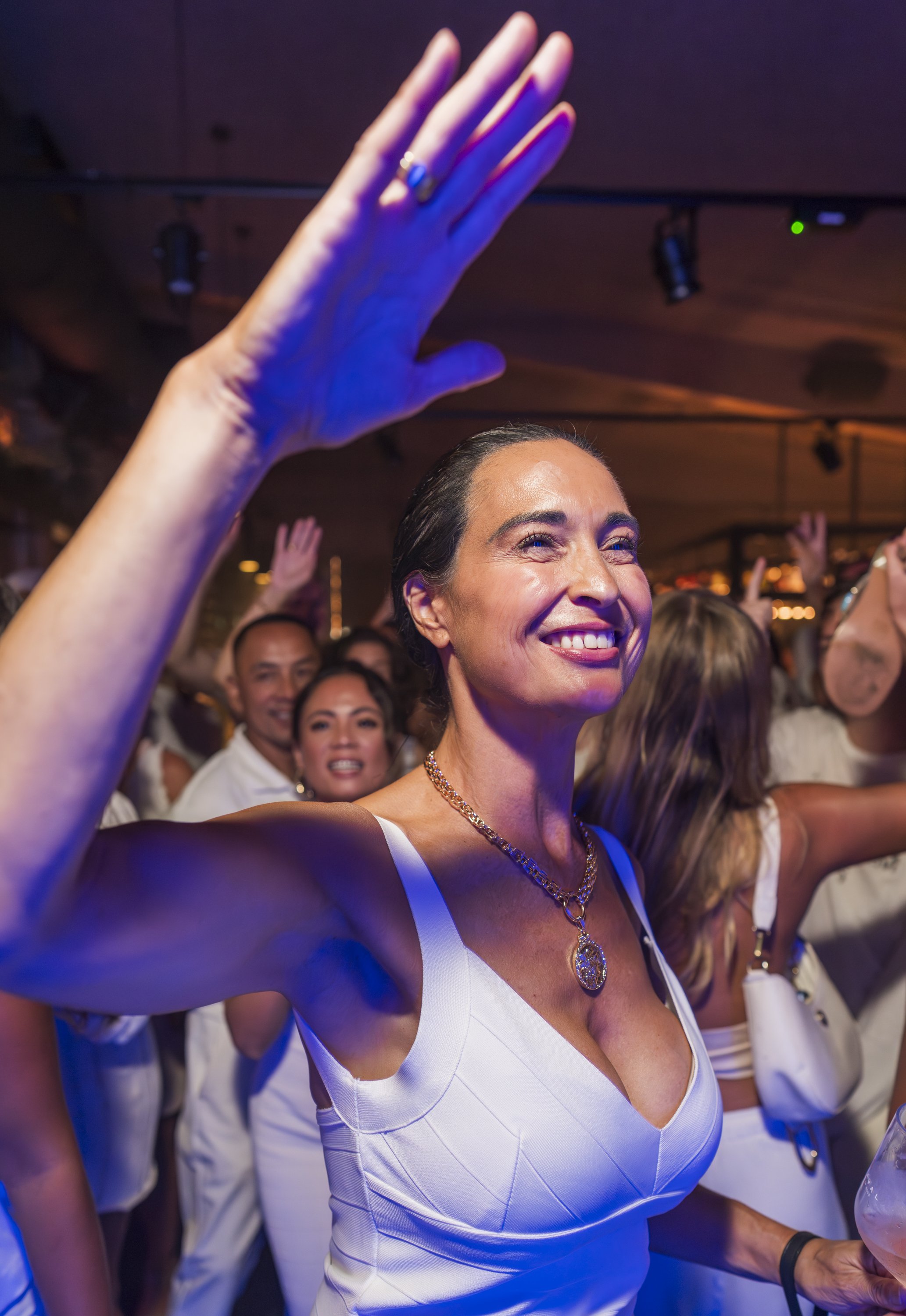 Een vrouw met een witte jurk en een ketting die op een feest of concert is, met andere mensen op de achtergrond, allemaal met glimlachen en in een feestelijke sfeer.