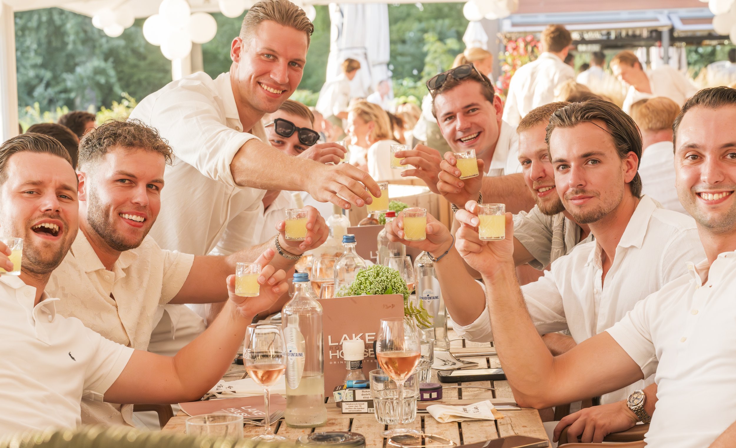 Een groep vrienden die samen plezier hebben bij een restaurant of café, zij heffen hun glazen in een toast, er is een gezellige sfeer met drukke achtergrond en groene omgeving.