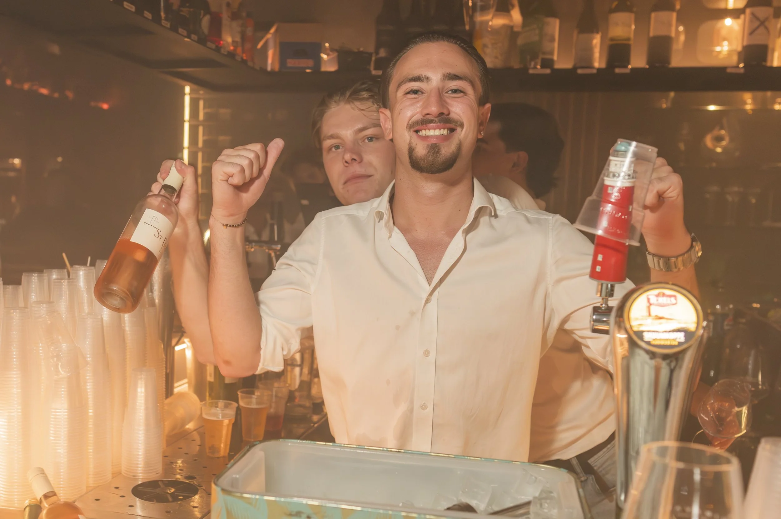 Man in witte shirt achter de bar met een brede glimlach, omringd door plastic bekers, flessen en barproducten in een bar of clubomgeving.