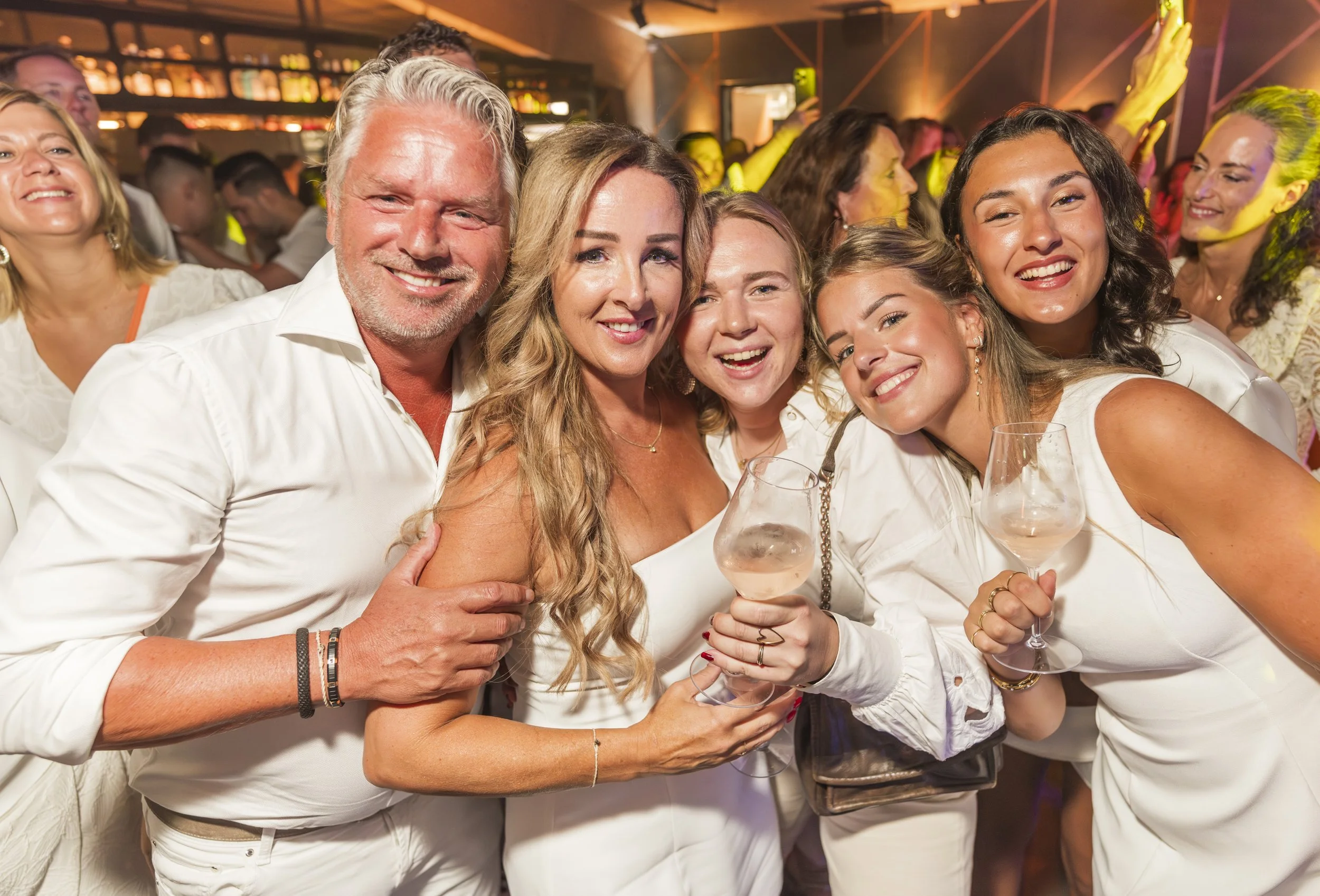 Vijf mensen die samen poseren tijdens een feest, lachend en met glazen wijn in de hand, in een feestzaal met kleurrijk licht.