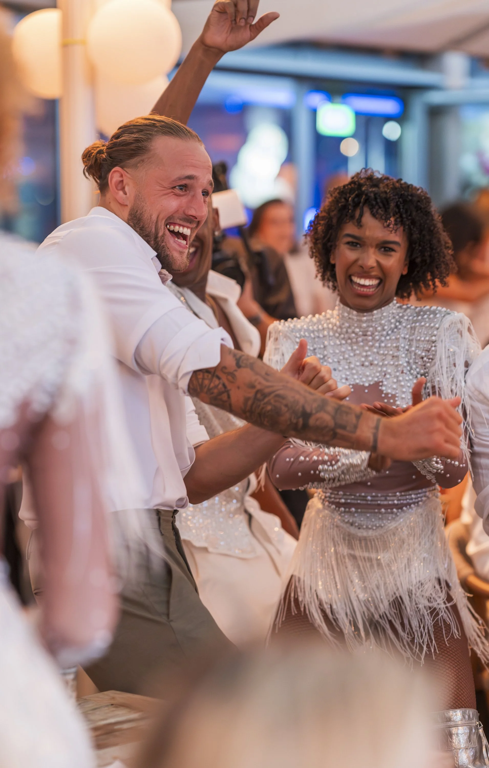 Mensen dansen en lachen tijdens een feest, de vrouw draagt een witte jurk met parels en franjes, de man heeft lang haar in een paardenstaart en tattoos op zijn armen.