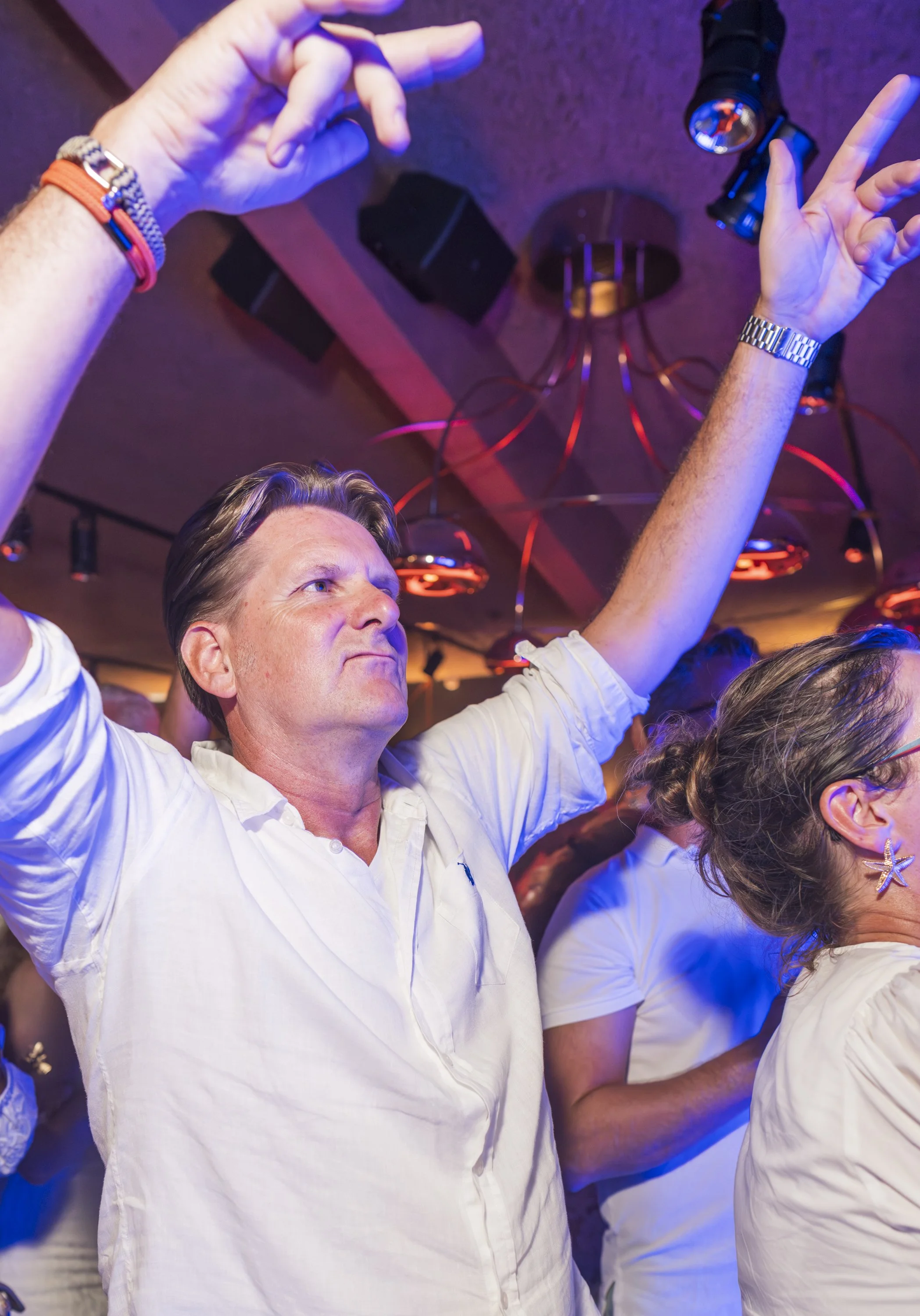 Man in witte blouse met armbanden en horloge heeft handen omhoog en geniet van muziek of dans op een feestje met blauwe en oranje verlichting.