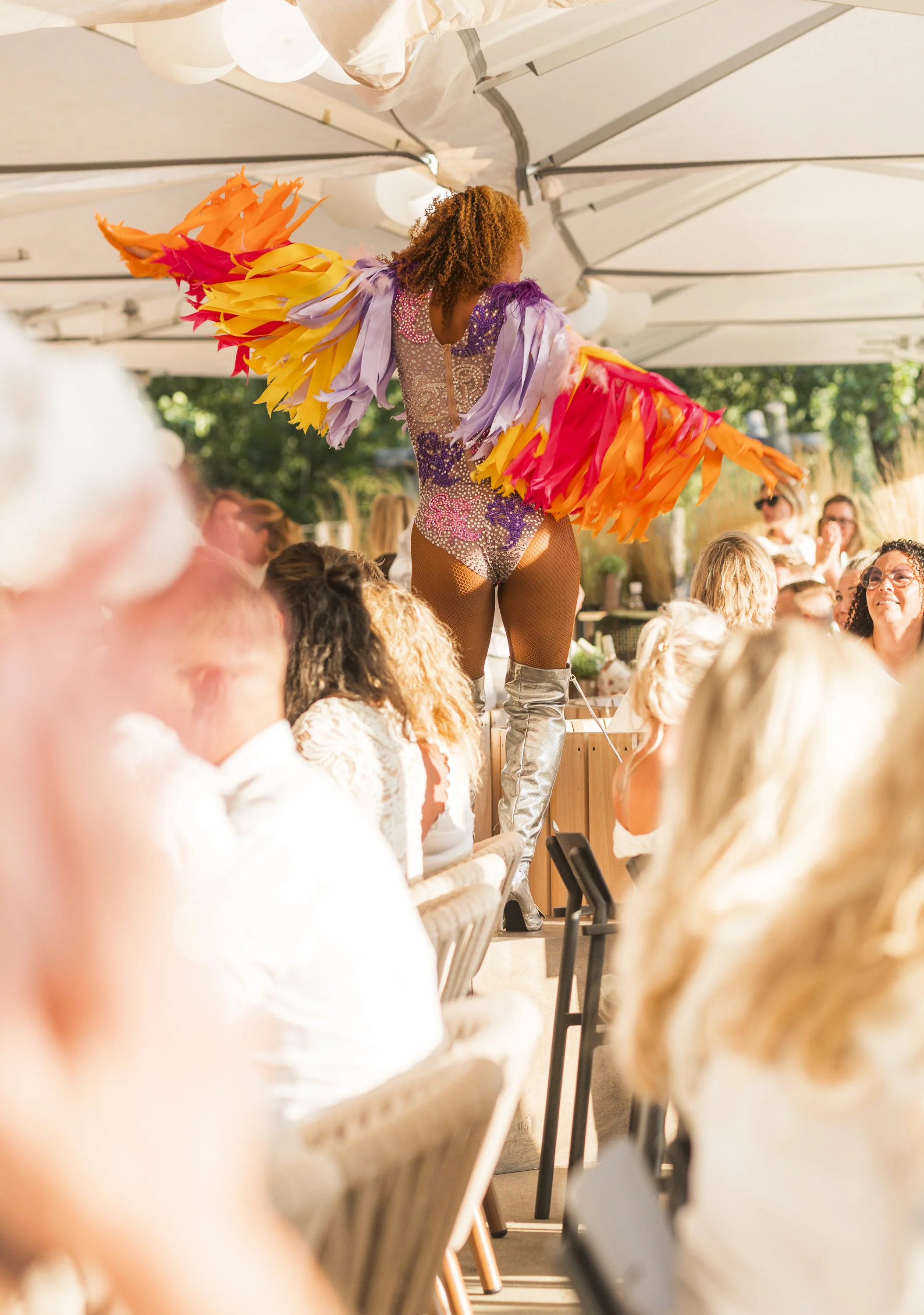 Een performer met kleurrijke, gelaagde vleugelachtige kostuum tijdens een show voor een publiek onder een grote overkapping.