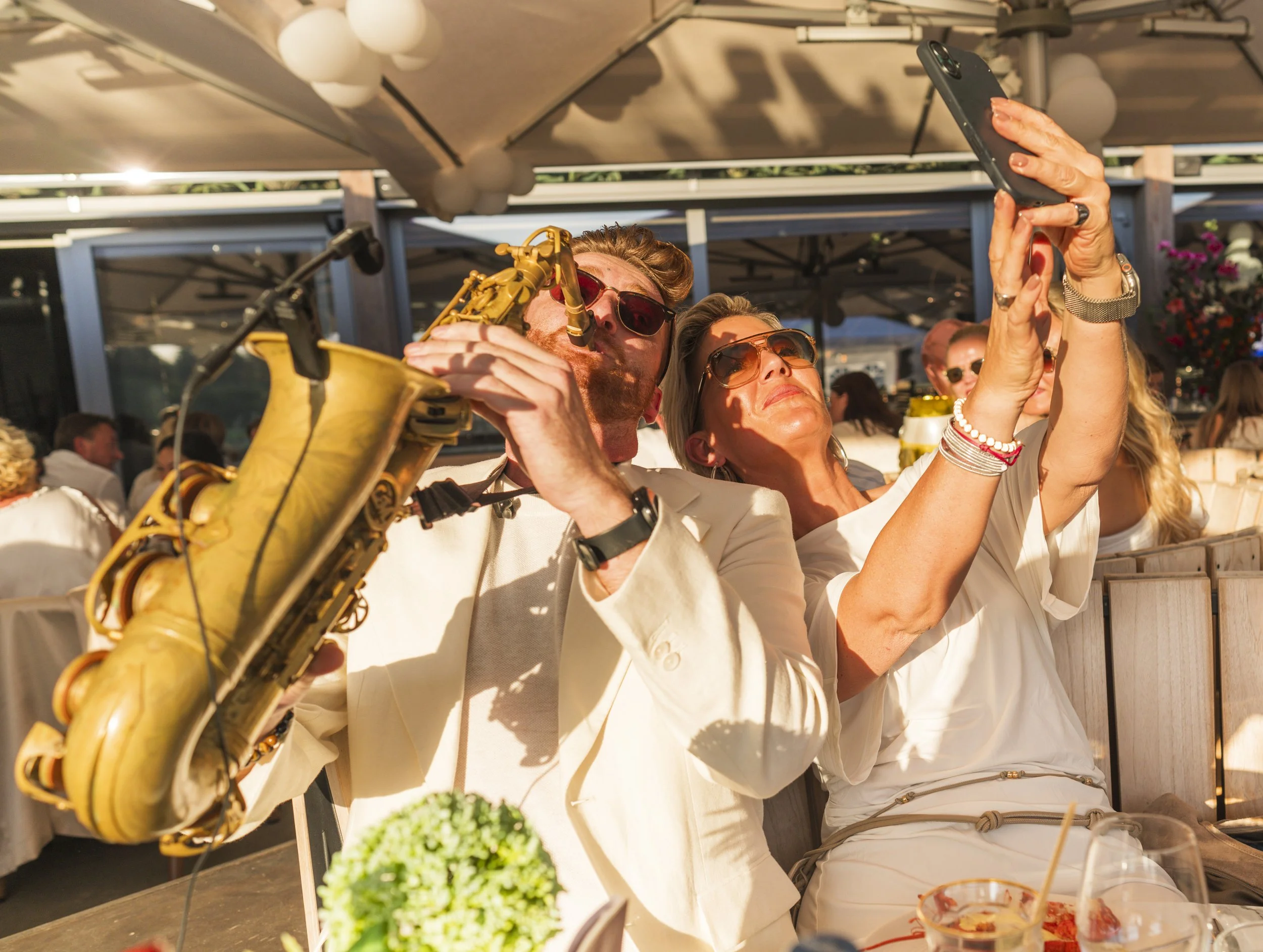 Drie mensen maken een selfie met een smartphone op een terras, omgeven door decoraties en andere mensen.