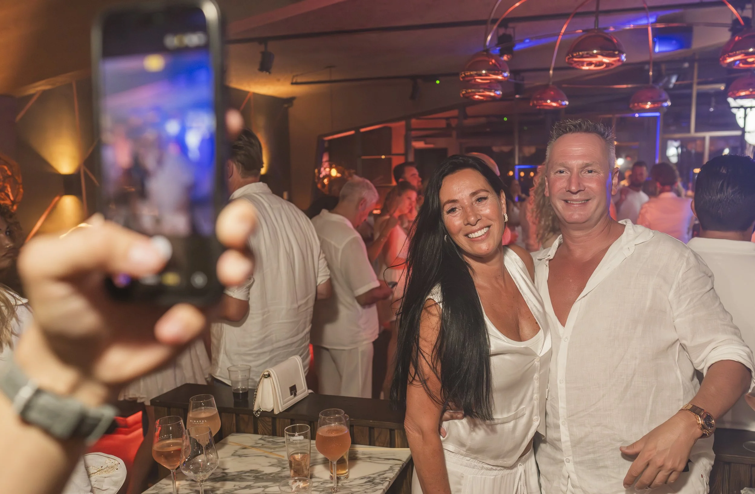 Een man en een vrouw poseren voor een foto in een feestzaal, omringd door andere feestvierders, met drankjes op een tafel daarnaast en kleurrijk verlicht decor.