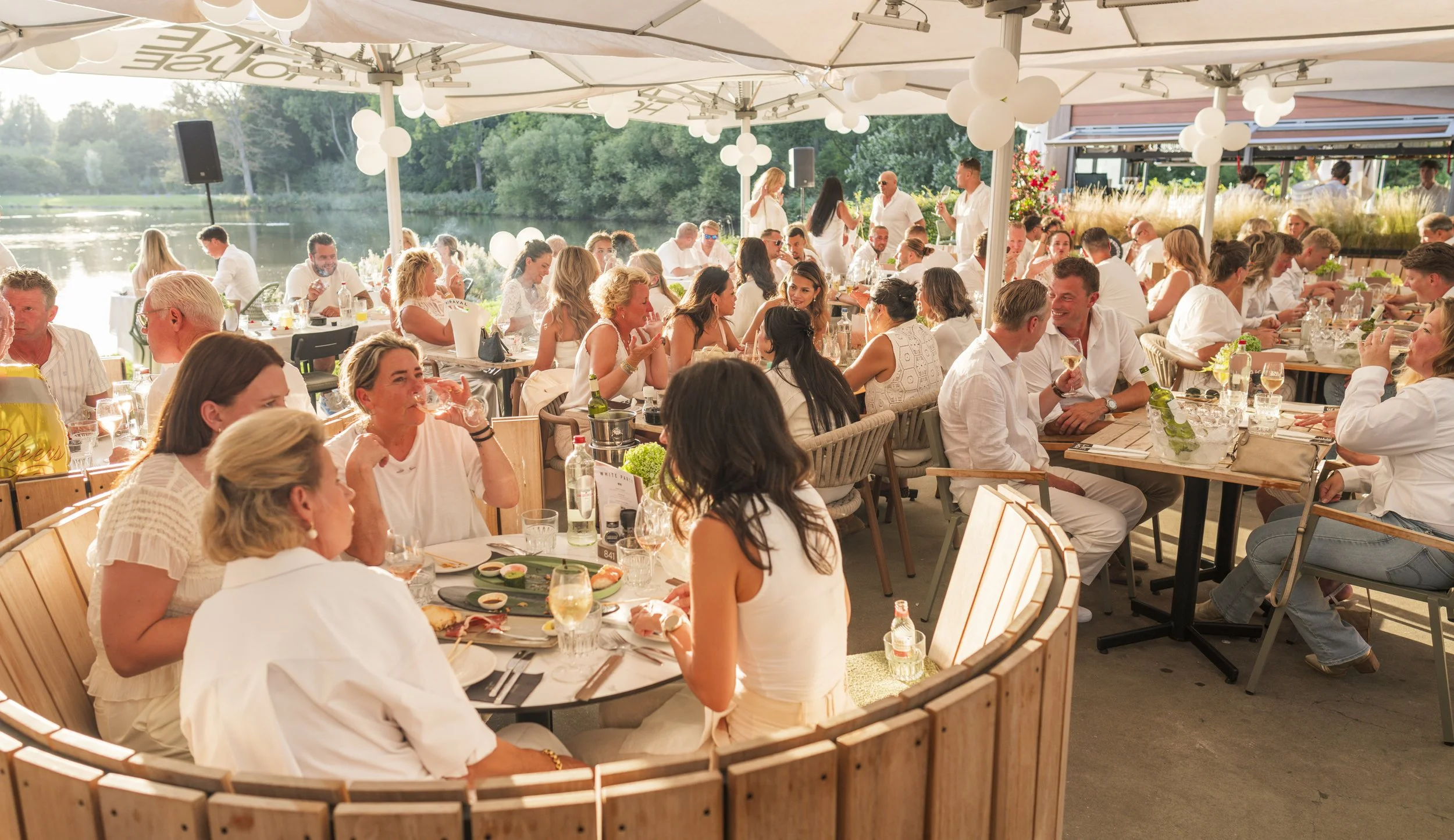 Gezellige groepsfoto van mensen die genieten van een diner samen, onder een parasol aan een rivier met groene omgeving op de achtergrond.