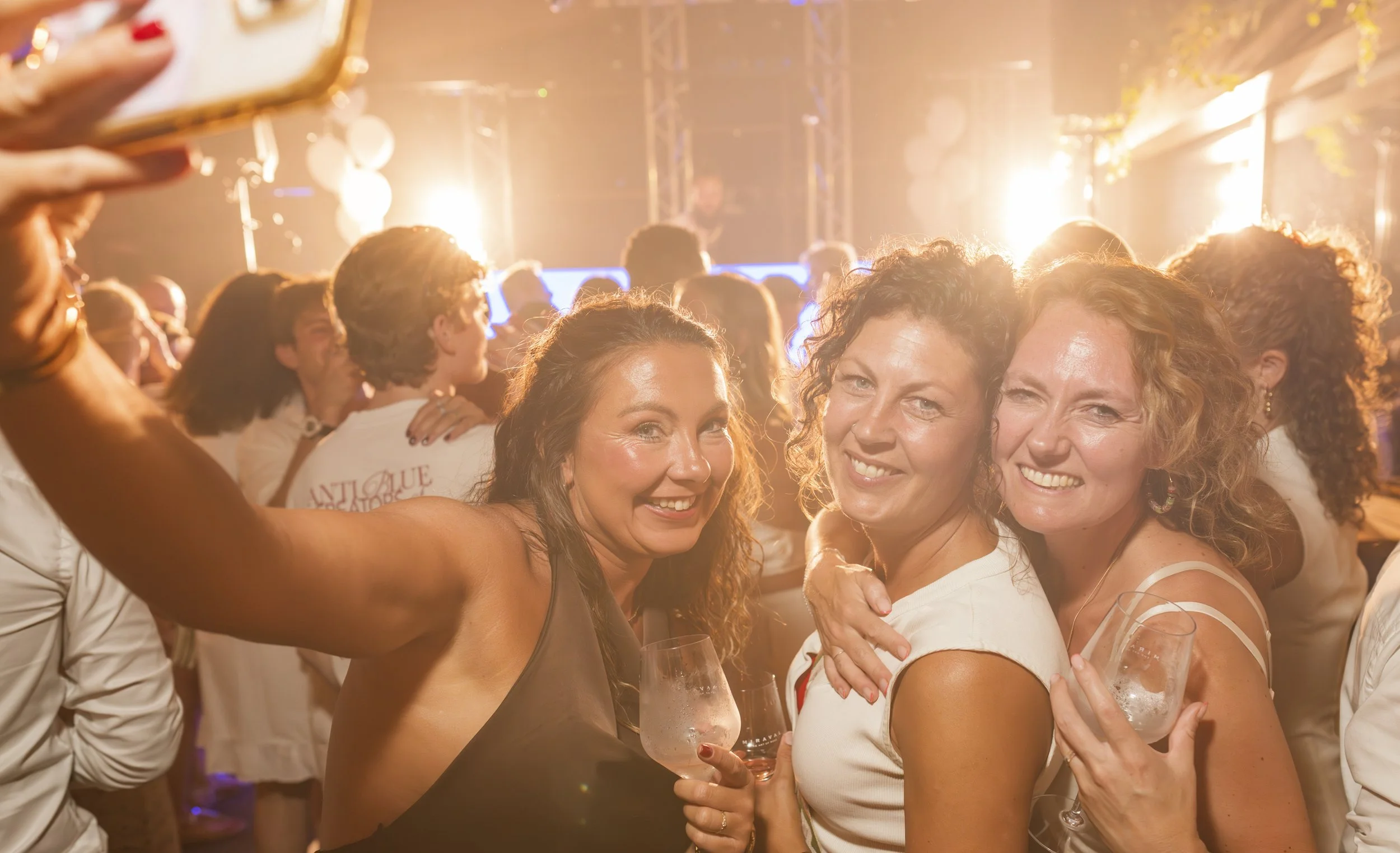 Vrouw die lacht en een glas wijn vasthoudt terwijl ze omarmd wordt door een andere vrouw op een feest of evenement met veel mensen en heldere verlichting.