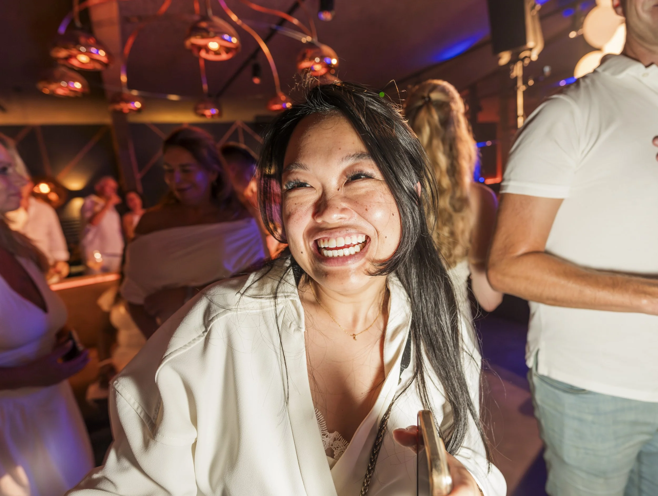 Een lachende vrouw met zwart haar die op een feestje of dansvloer staat, omringd door andere mensen, onder kleurrijk licht.