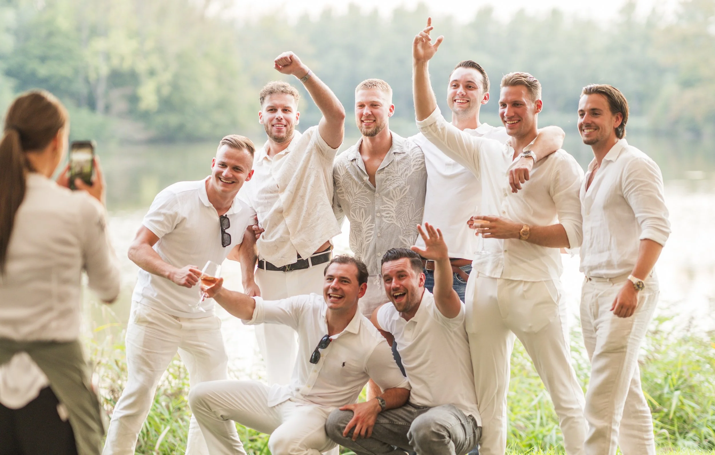 Groep mannen die lachen en poseren voor een foto bij een waterside, ze dragen allemaal witte kleding, een vrouw neemt een foto van ze met haar mobiele telefoon.