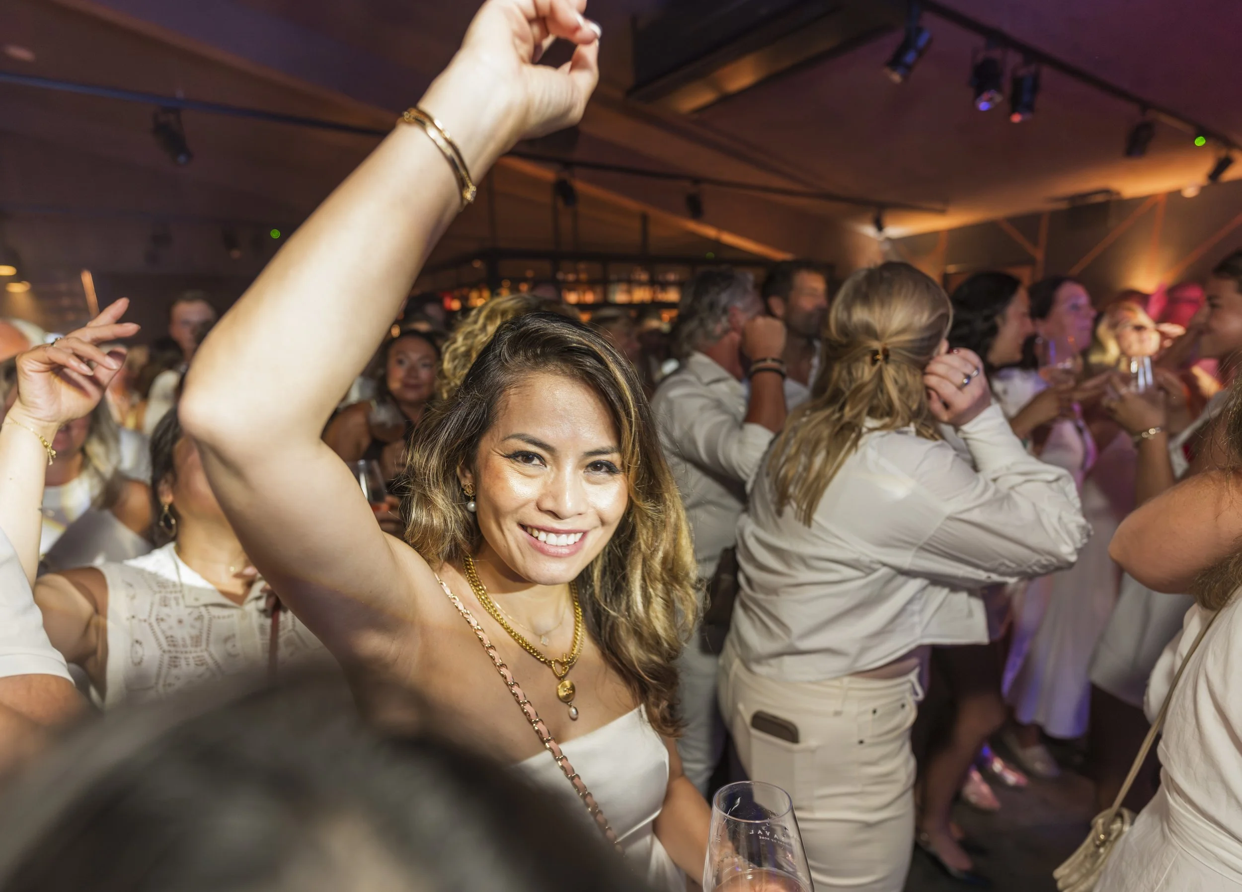 Vrolijk uitziende vrouw met goudkleurige kettingen en armbanden, omringd door mensen die praten en lachen tijdens een feestje of evenement.