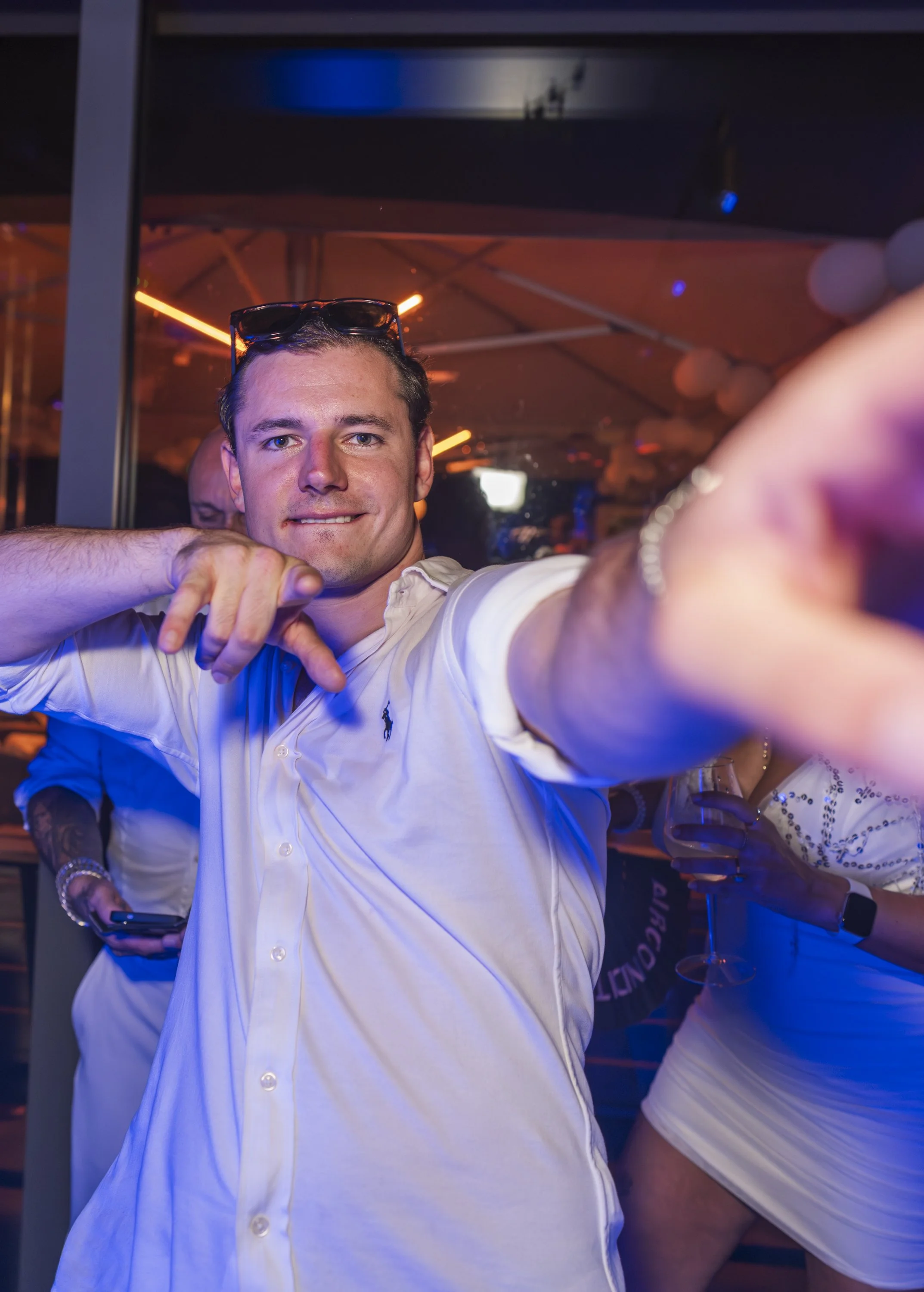 Een man die een selfie neemt bij een feest of club, met een glas wijn in de hand en een neonverlichting op de achtergrond, in een feestelijke sfeer.