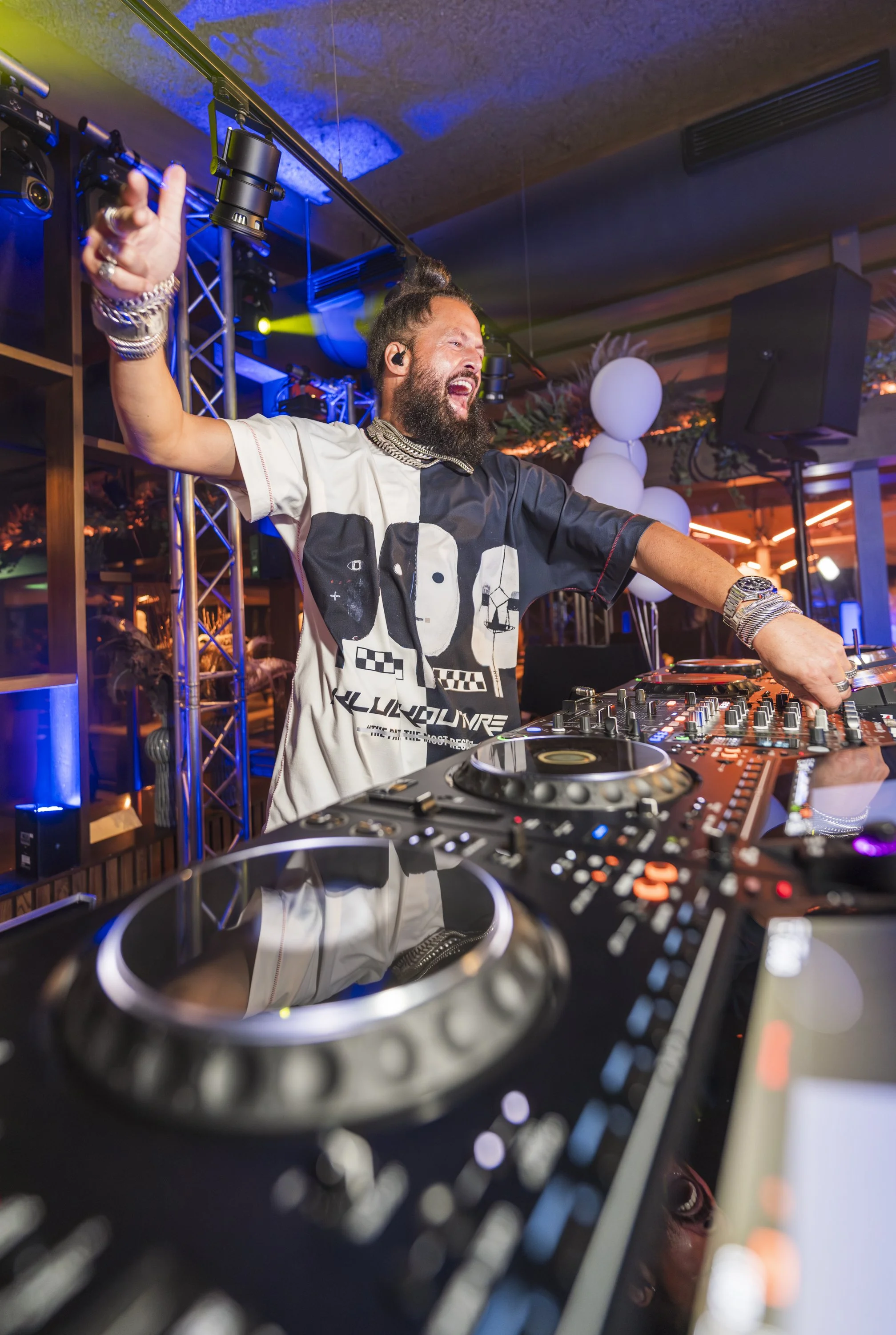 Een DJ met een microfoon en koptelefoon achter een draaitafel tijdens een feestje, met kleurrijk licht en decoraties op de achtergrond.