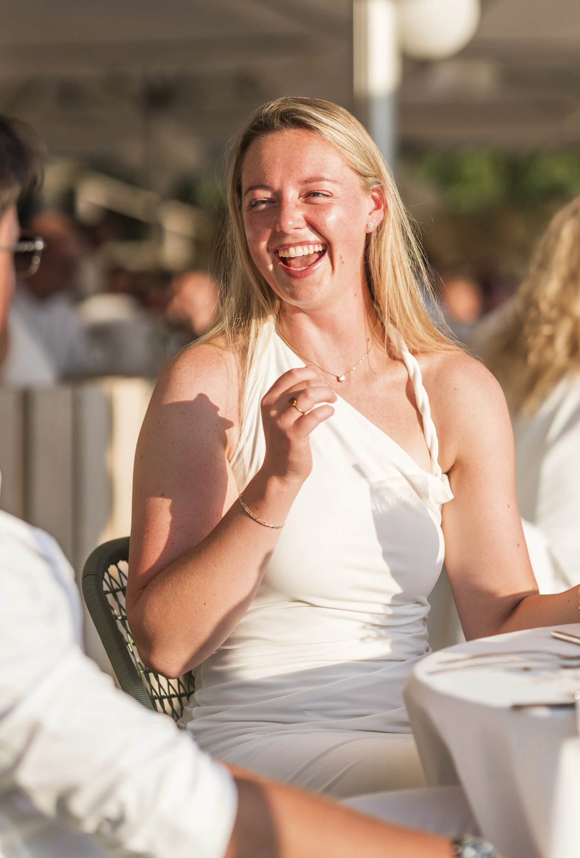 Een lachende jonge vrouw met blond lang haar draagt een witte jurk en is te zien tijdens een feest of bruiloft, met anderen op de achtergrond.