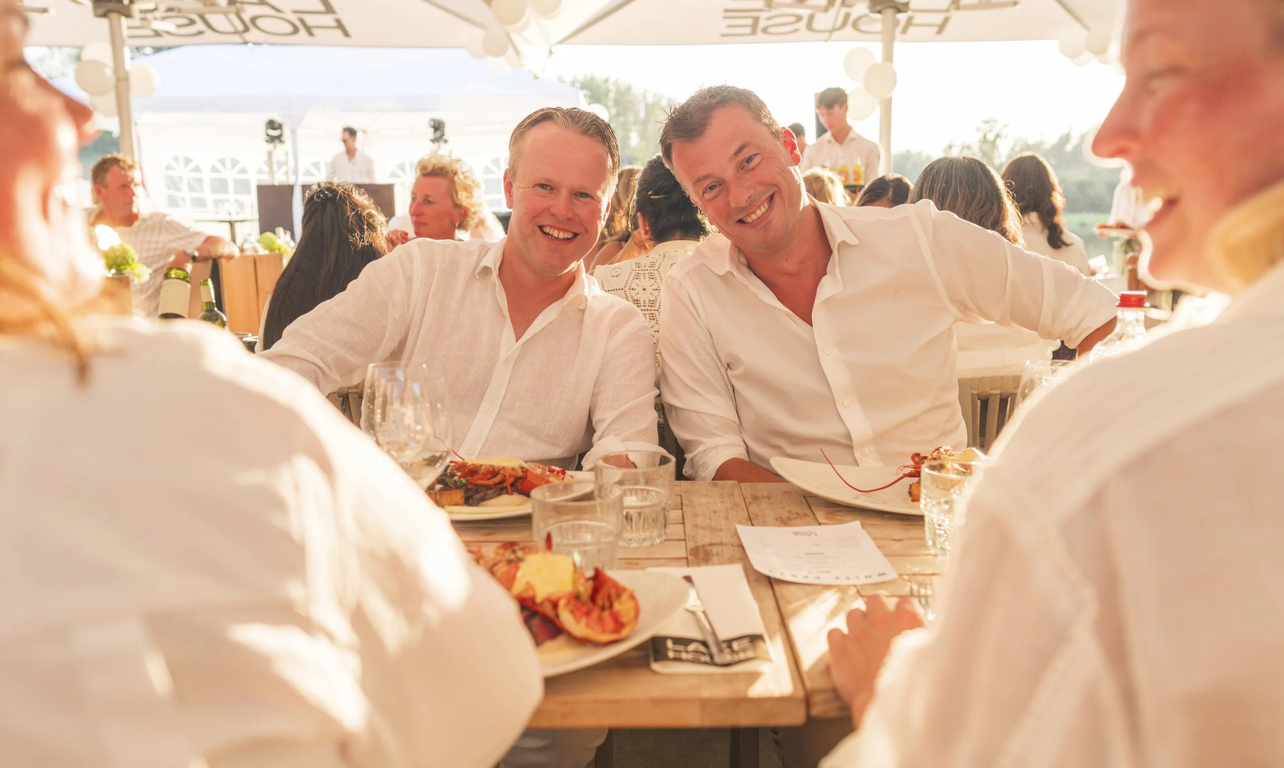 Twee lachende mannen zitten aan een tafel tijdens een buitendiner, omgeven door andere gasten en een feesttent met witte ballonnen in de achtergrond. Ze dragen witte overhemden en genieten van de sfeer.