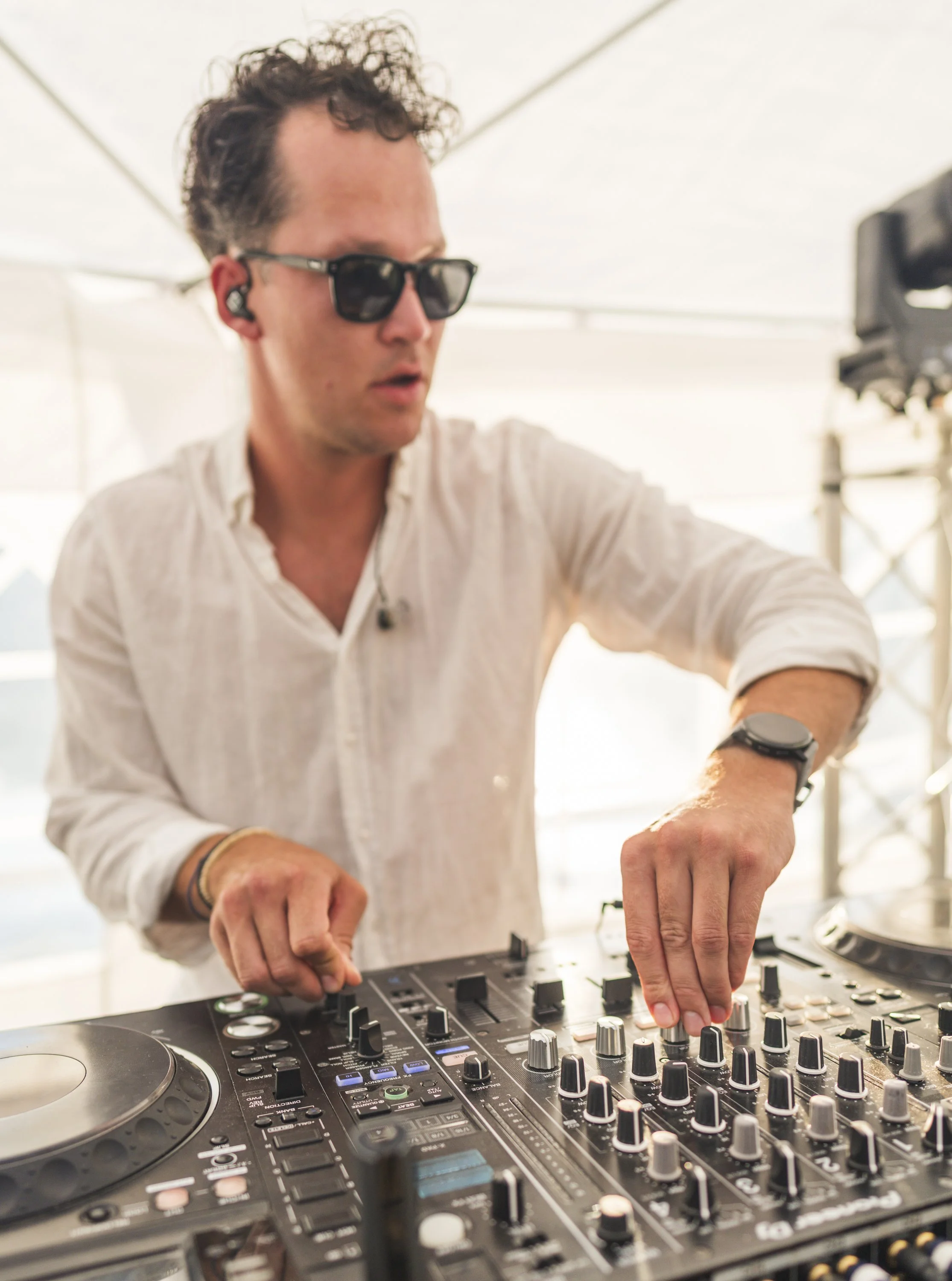 Man met donker haar, zonnebril en witte blouse die electronic muziek draait op DJ-set, binnen in een lichte ruimte.