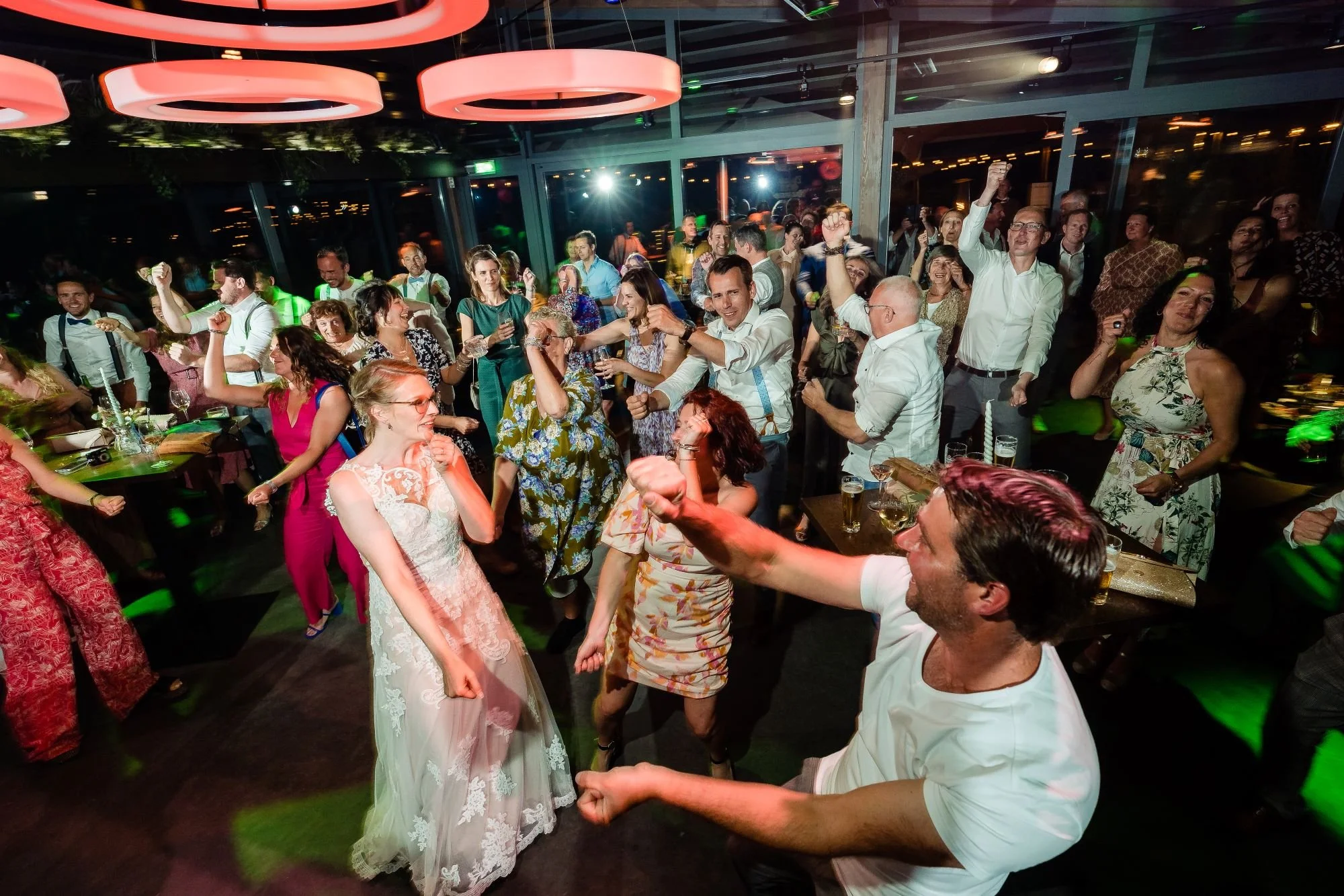 People dancing and celebrating at a lively party celebration in a modern venue with pink ceiling lights and large windows.