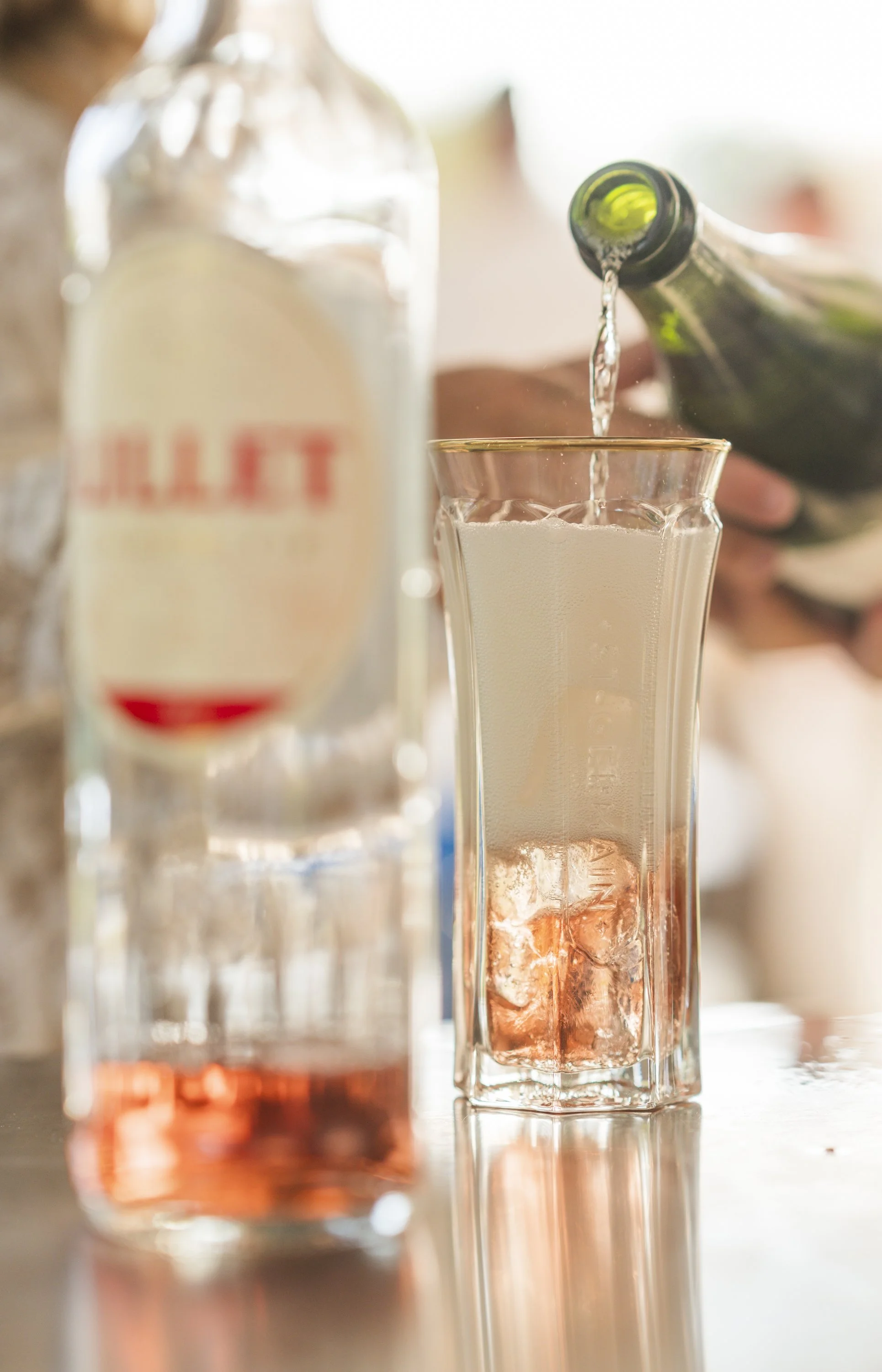 Een glas met een lichtroze drankje en ijsblokjes, terwijl iemand een fles champagne inschenkt.
