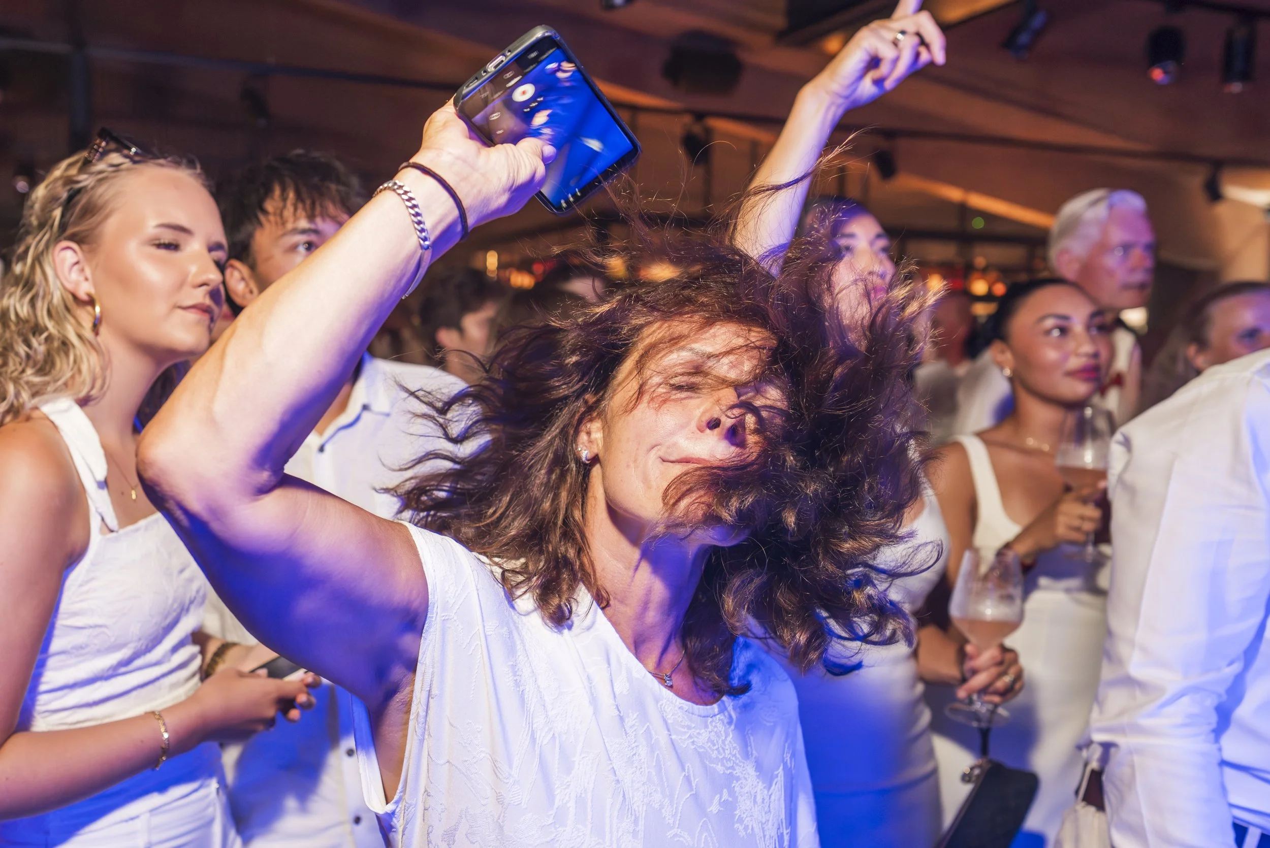 Een groep mensen op een feest of concert, sommigen maken foto's of video's met hun telefoons, terwijl anderen dansen en genieten.