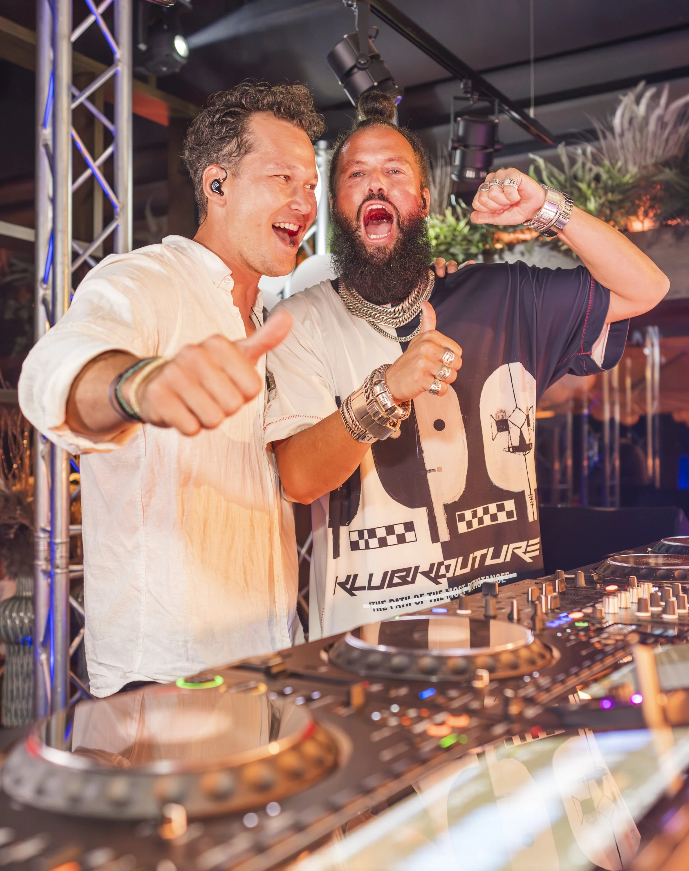 Twee mannen die samen plezier maken met dj-gear, een van hen draagt een zwart shirt met tekst en sieraden, de andere draagt een wit overhemd en een in-ear monitor.
