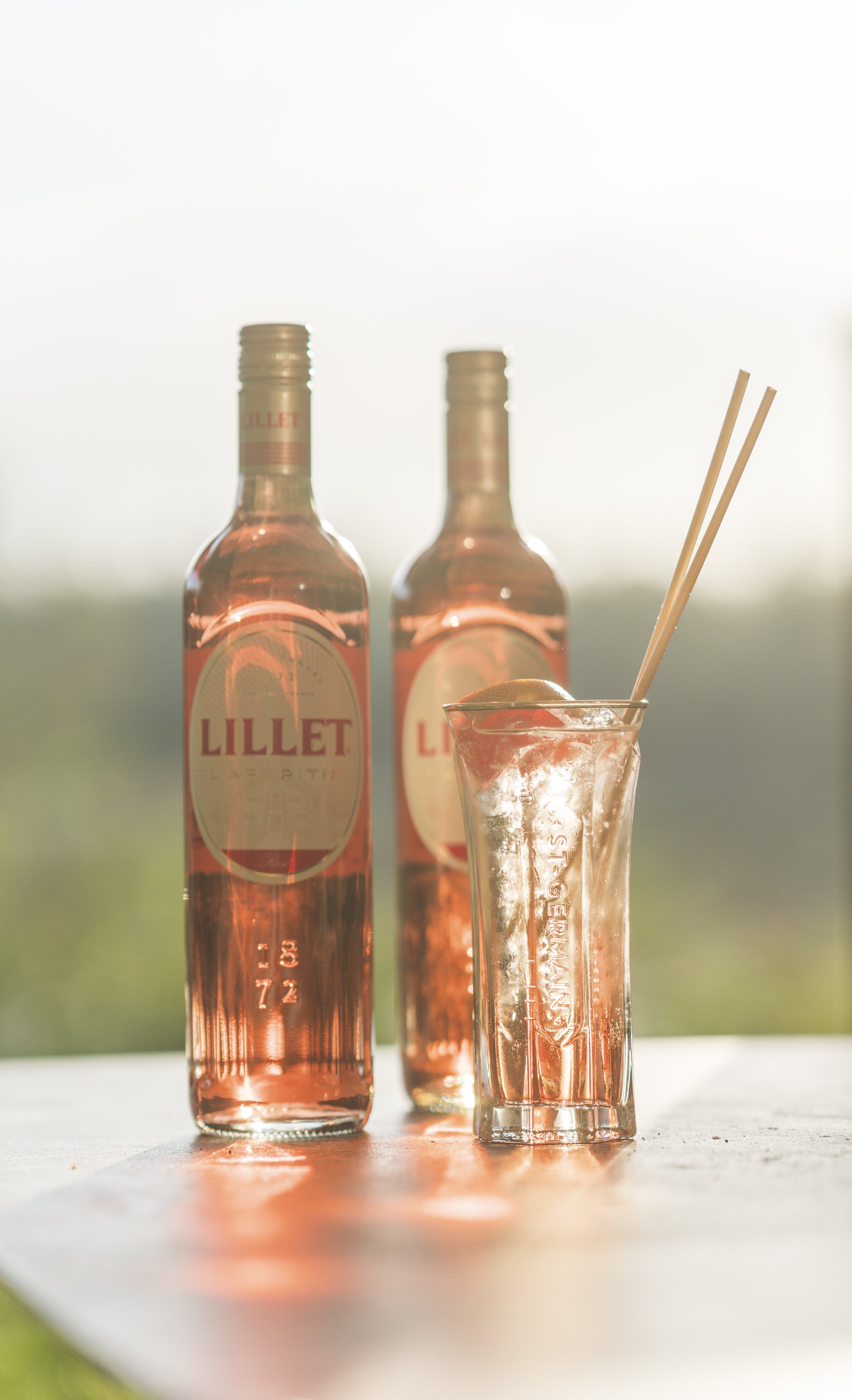 Twee flesjes rosé wijn en een glas met ijs en twee rietjes op een lichte tafel, met een groene buitenachtergrond.