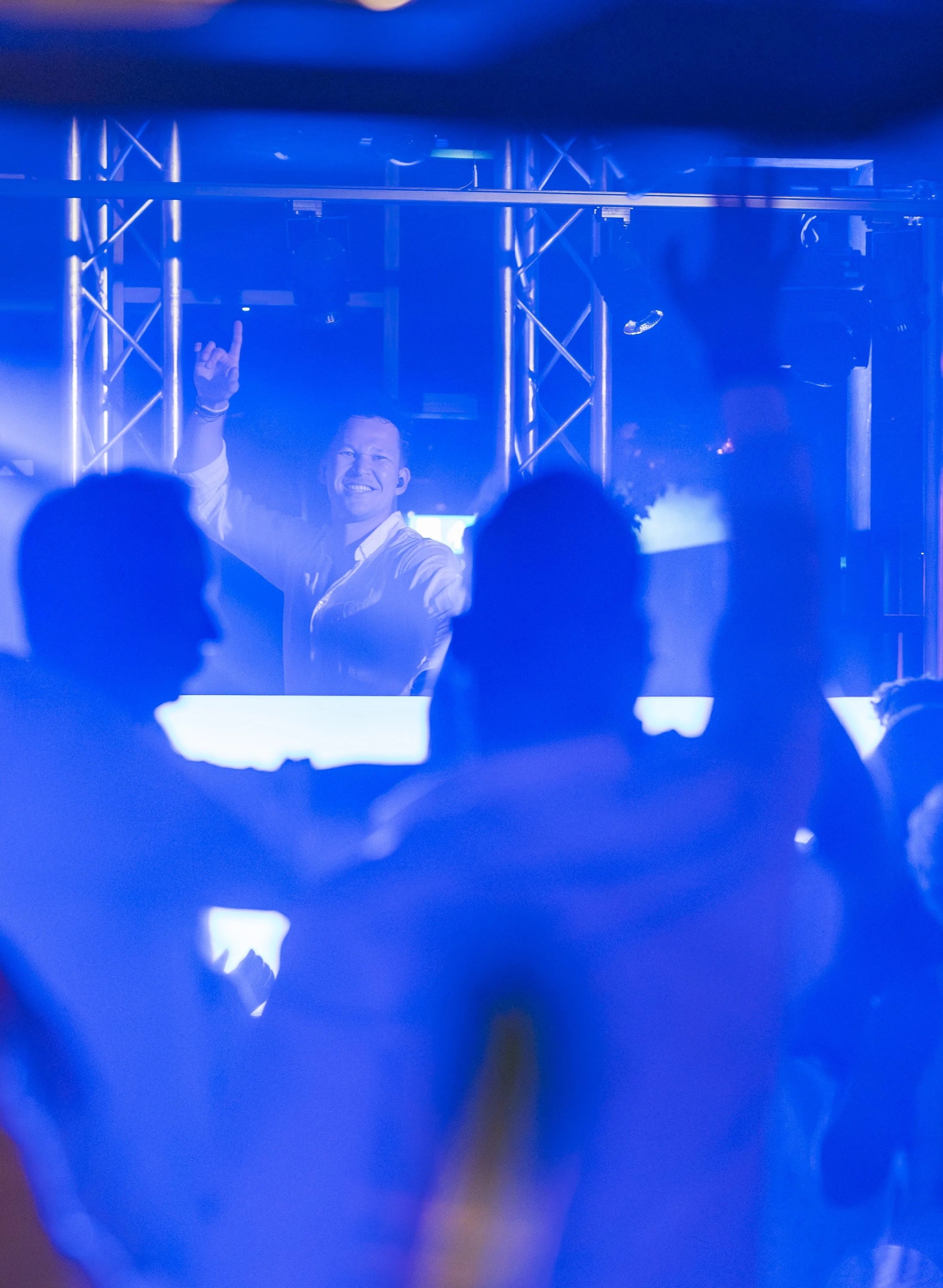 Een feestelijke scène met een lachende man die opstaat en de hand omhoog steekt, gefilmd door een blauw getint glas of een blauw filtreerfilter in een club of op een feest, met silhouetten van andere mensen op de achtergrond.