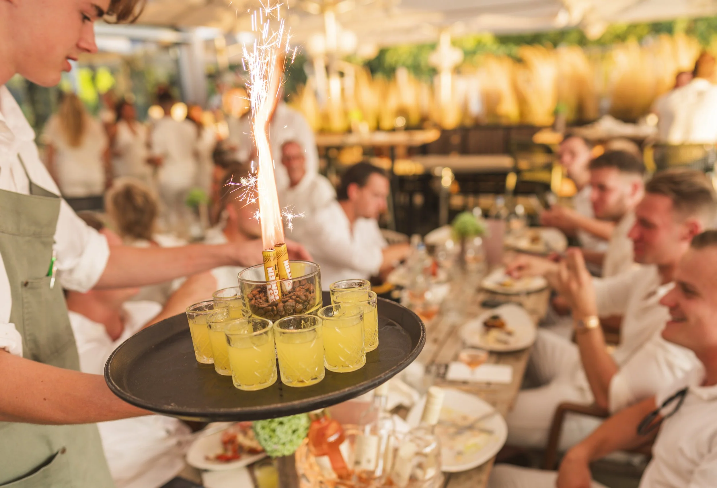 Een ober brengt een dienblad met shotjes heldergelucht drankje met een vuurwerk-fakkel naar een groep mensen die aan een tafel zitten, in een gezellige, feestelijke omgeving.
