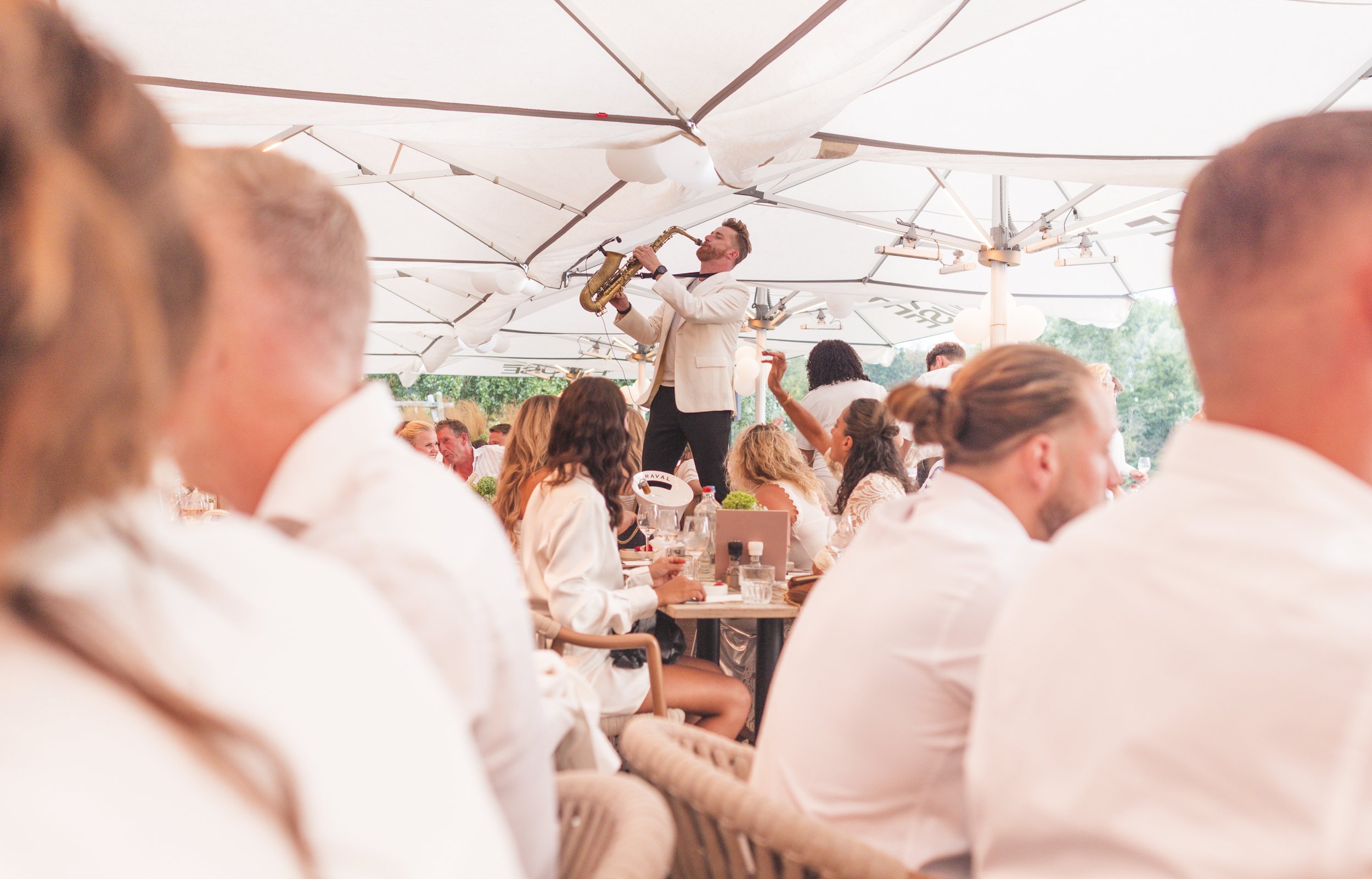 Man speelt saxofoon onder een grote witte parasol op een buitenevenement, omringd door mensen die aan tafels zitten.
