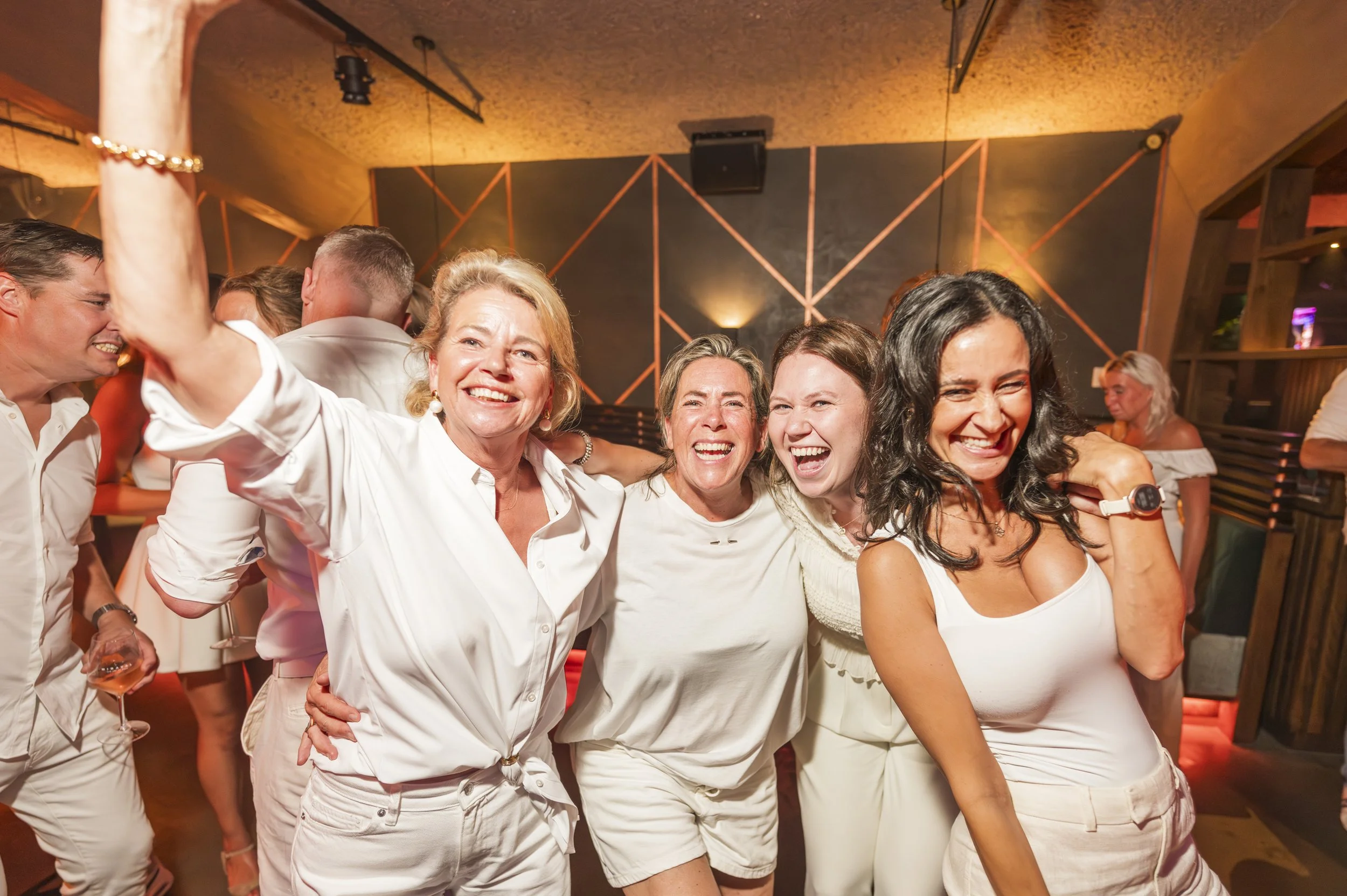 Groep mensen die samen lachen en plezier hebben op een feestje, met een warme ambiance en kleurrijke verlichting op de achtergrond.
